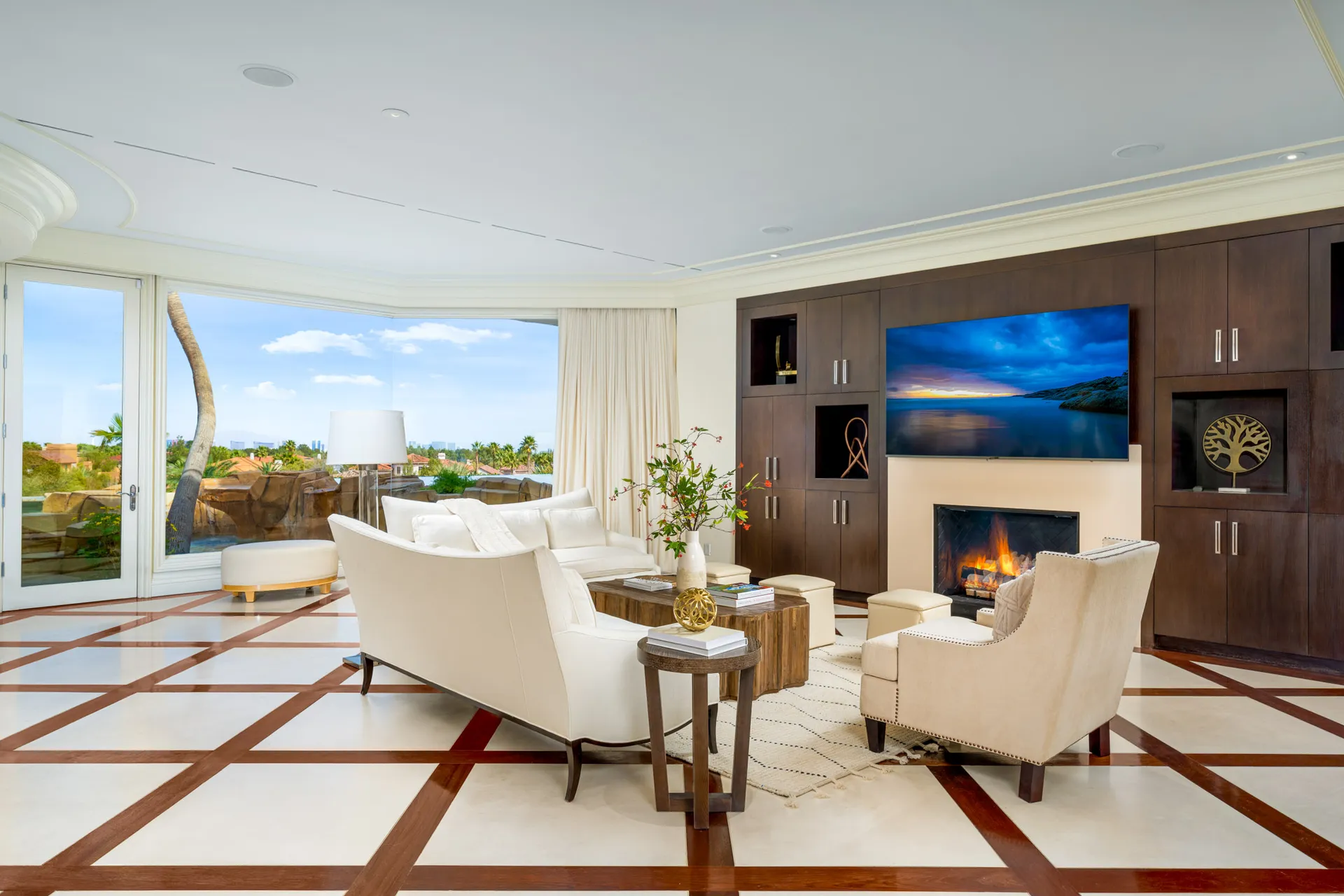 A living room with large glass windows overlooking an outdoor view, featuring a white sofa, a wooden coffee table, and a built-in fireplace with a TV above.