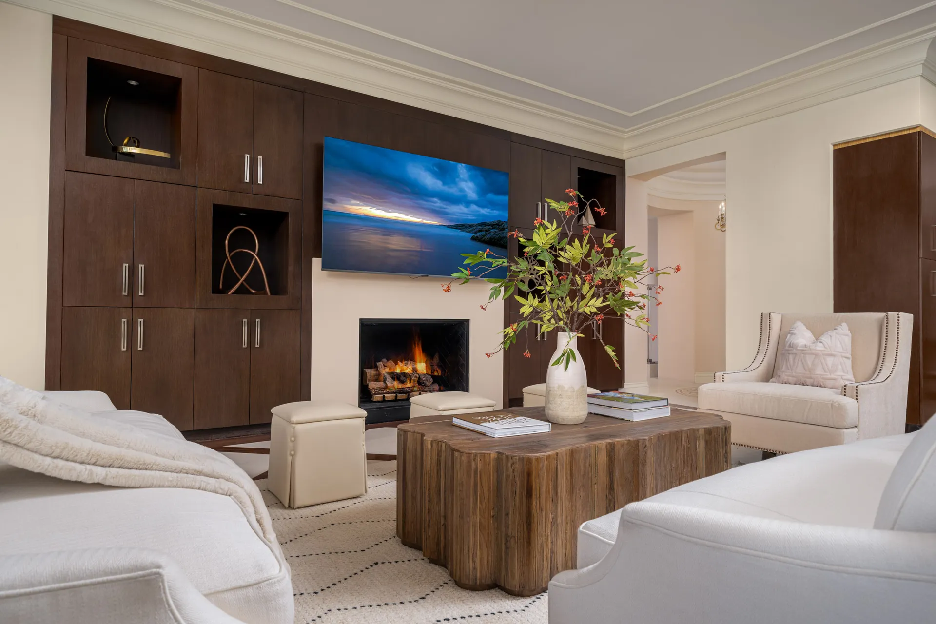 A living area featuring a built-in dark wood media unit with a TV mounted above a fireplace, a rustic wood coffee table with a vase, and neutral seating.