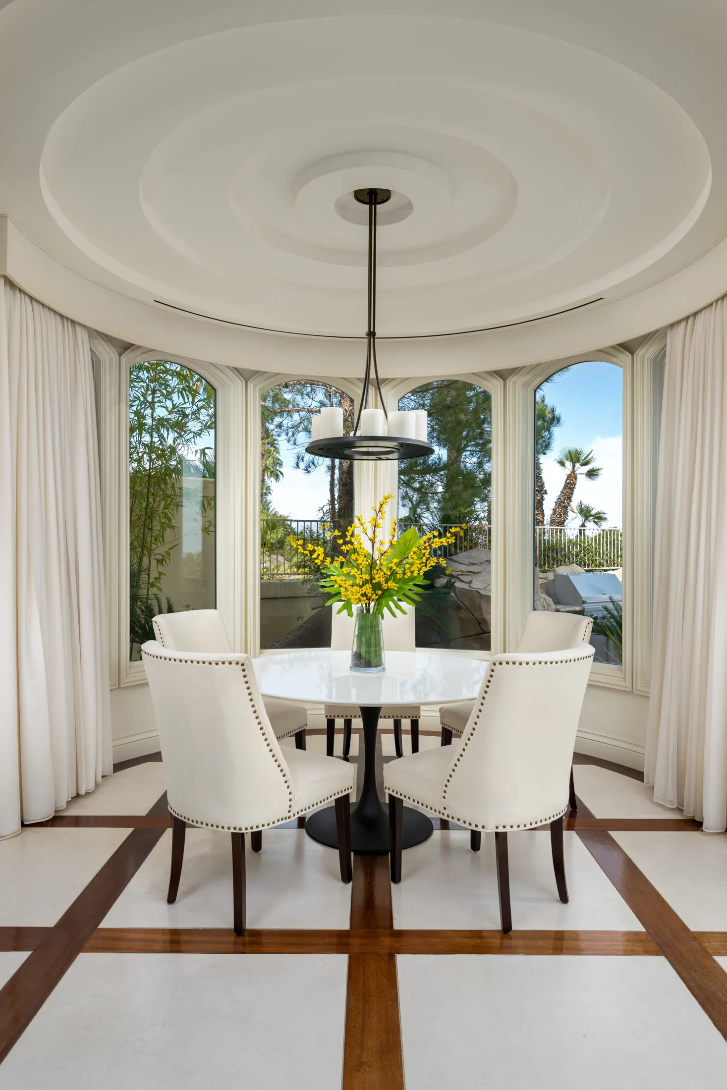A bright, circular dining nook with four white chairs around a round table. A simple chandelier hangs from the ceiling, and large windows offer garden views.