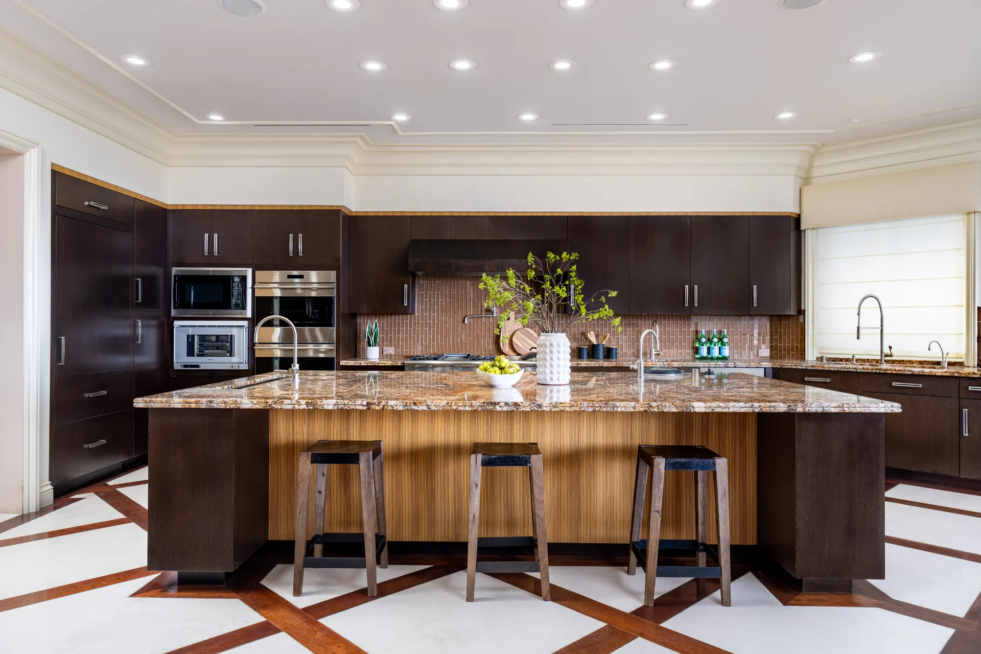 A spacious kitchen featuring dark wood cabinets, built-in appliances, and a large granite island. Three wooden barstools sit along the island’s edge.
