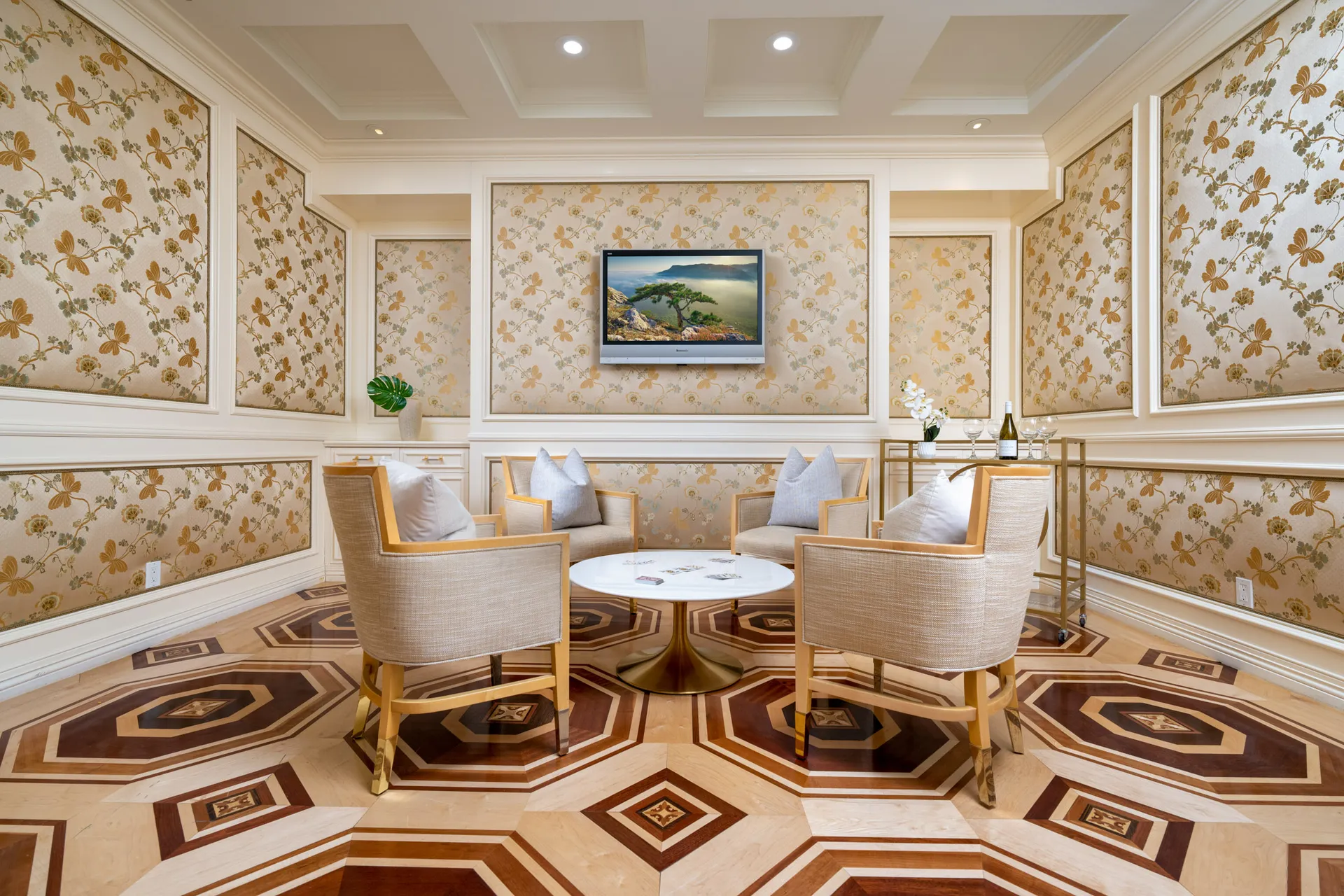 A sitting area with four light-toned chairs around a circular table, set against ornate floral wall panels. A bar cart stands nearby, and the inlaid wood floor features a striking hexagonal pattern.