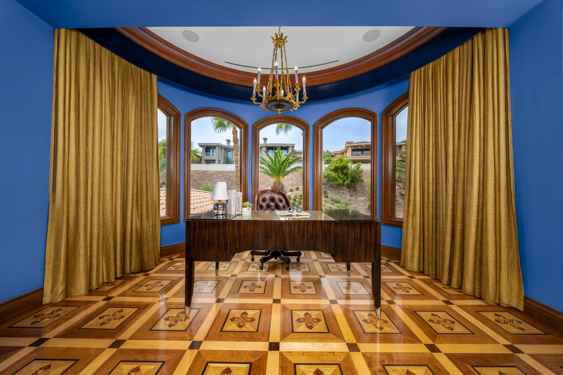 An elegant office with a curved wall of arched windows behind a polished wood desk and a tufted leather chair. Gold drapes and inlaid wood floors complete the luxurious look.