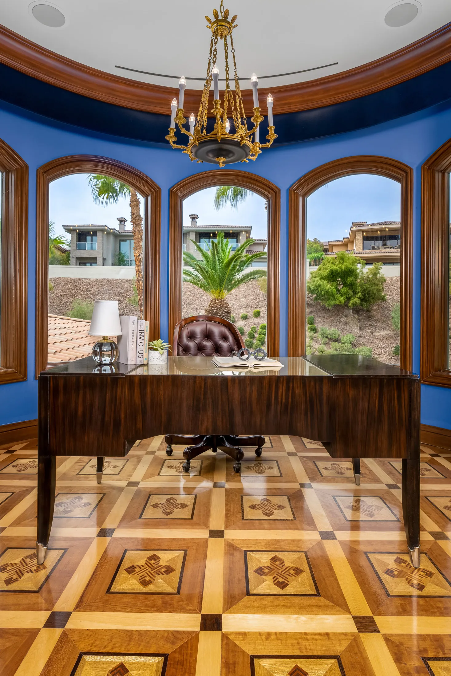 A curved wall of tall arched windows behind a polished wood desk with a tufted leather chair. An ornate chandelier and inlaid wood floor add a luxurious touch.