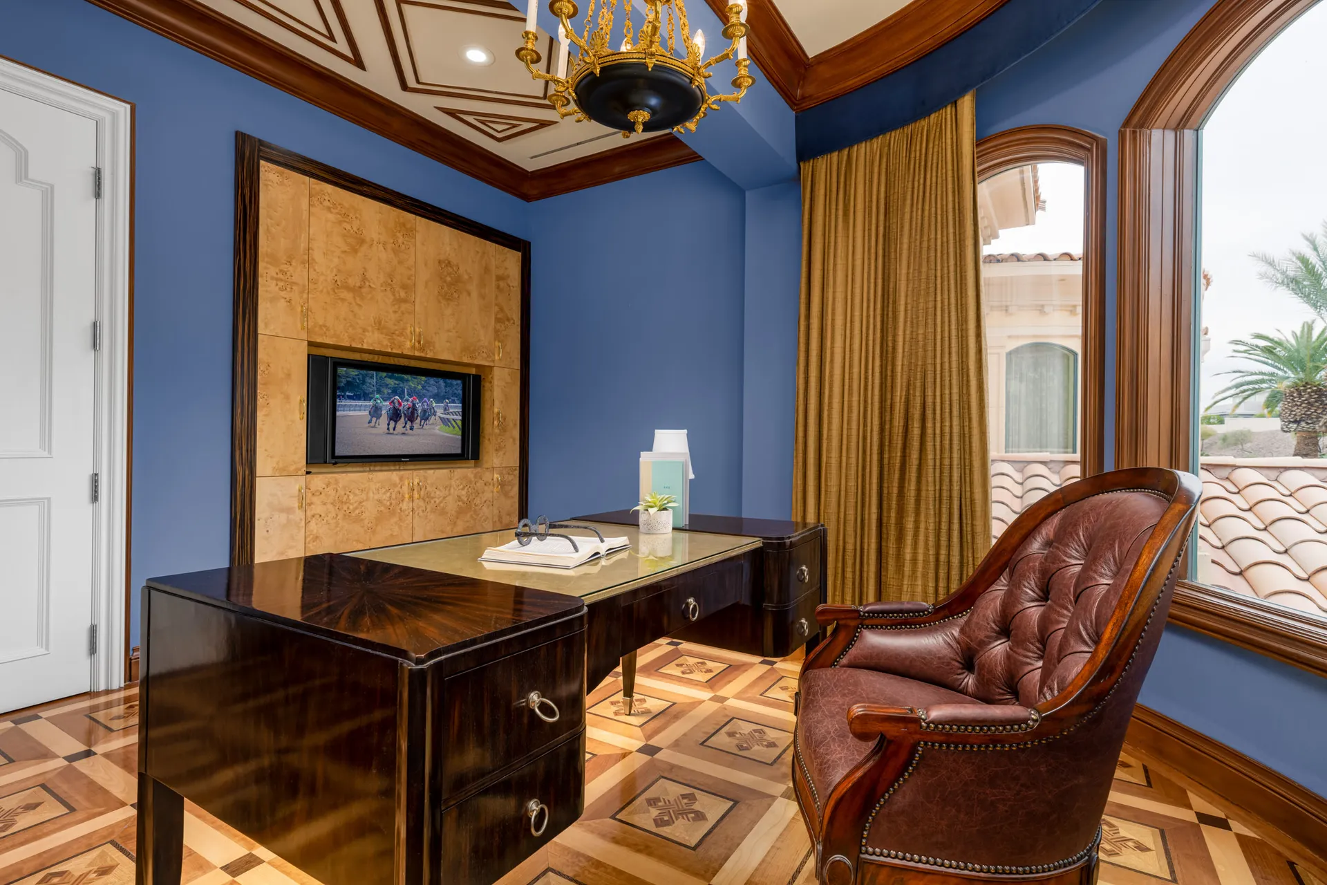 A home office with blue walls, an inlaid wood desk and floor, a built-in TV in a light wood cabinet, and a tufted leather chair. A gold chandelier hangs from the ornate ceiling.