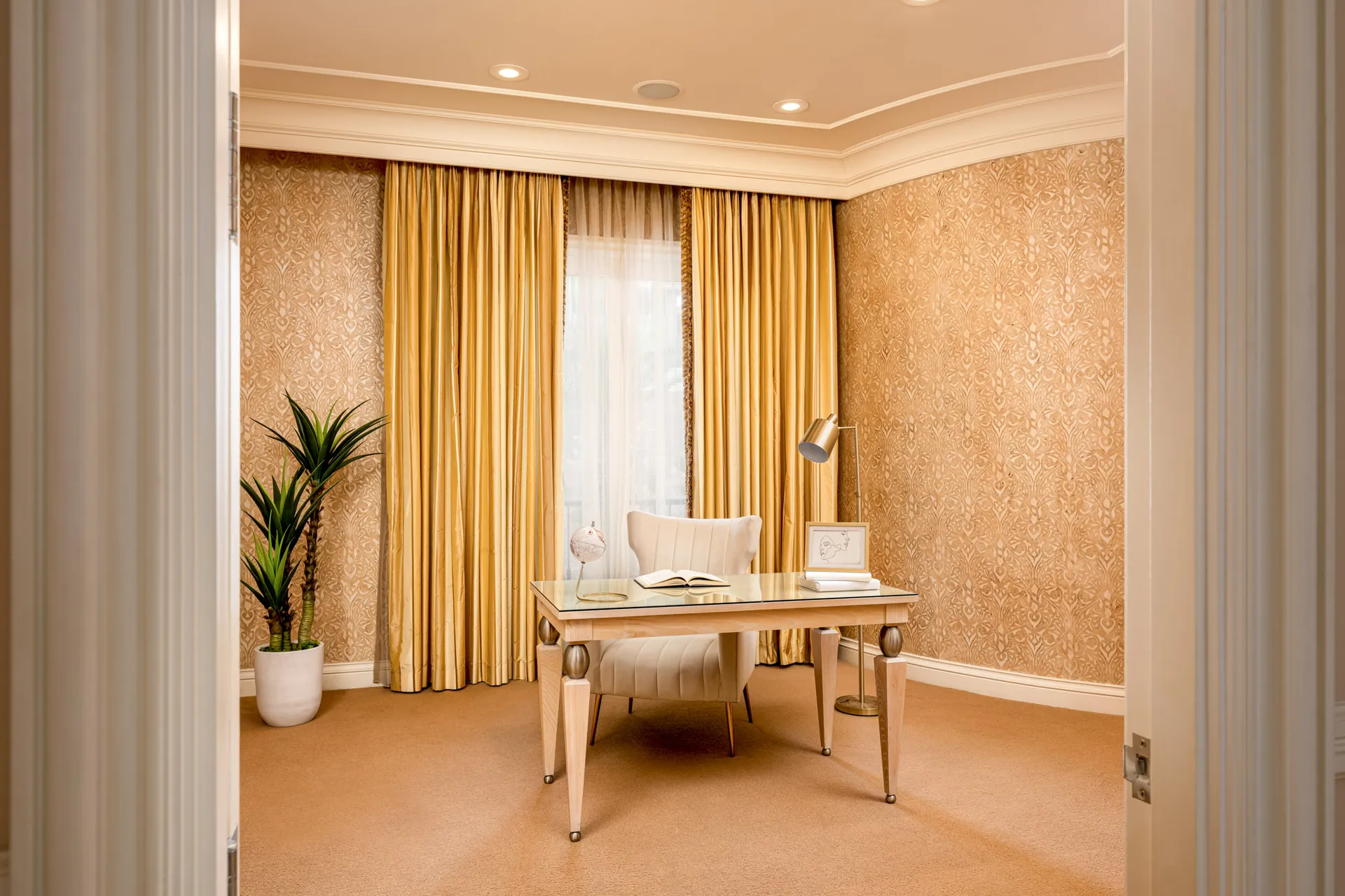 A warmly hued office featuring tall gold drapes and coordinating wallpaper. A modern wood desk with a tufted chair and a potted plant complete the space.