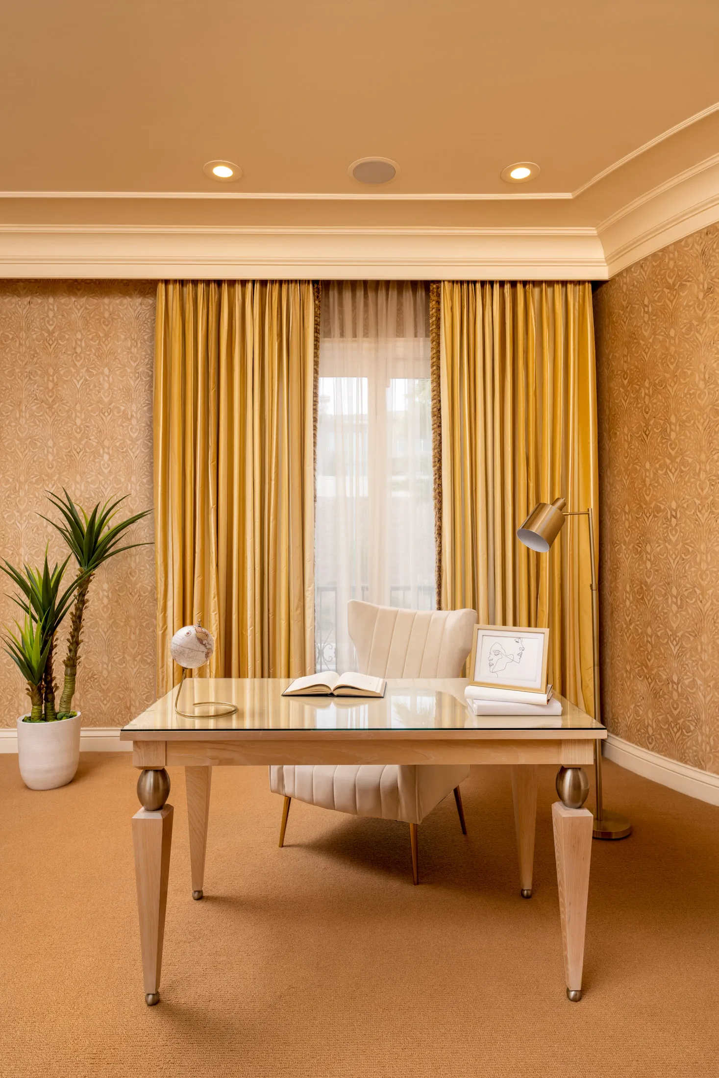 A warm home office with tall golden drapes, a light wood desk topped with a glass surface, and a tufted white chair. A small globe and framed artwork adorn the desk.