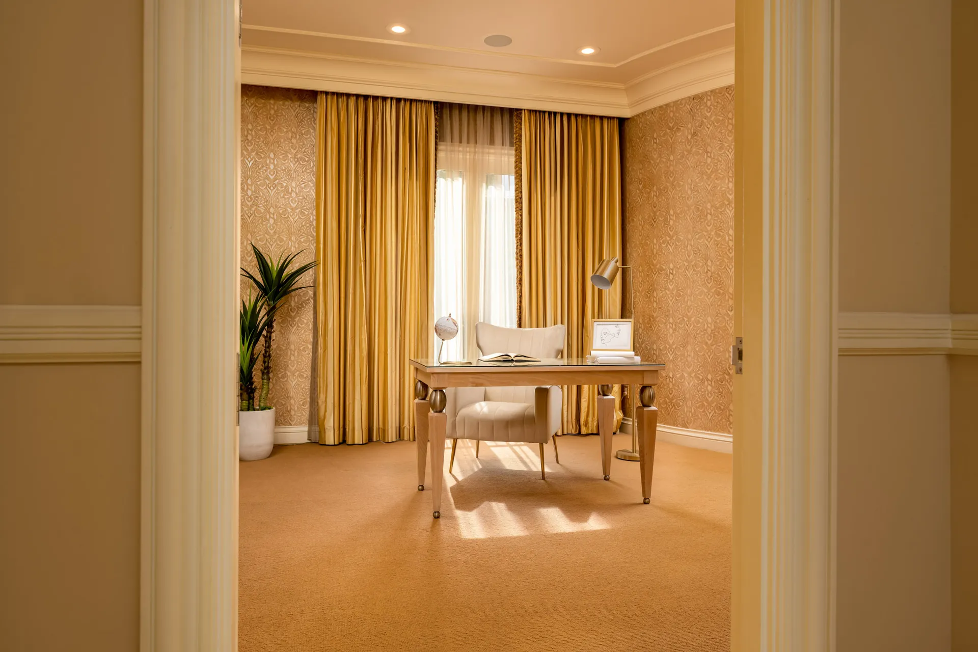 An elegant office with a light wood desk, plush chair, and gold drapery in front of tall windows. A potted plant stands to the left, complementing the warm decor.