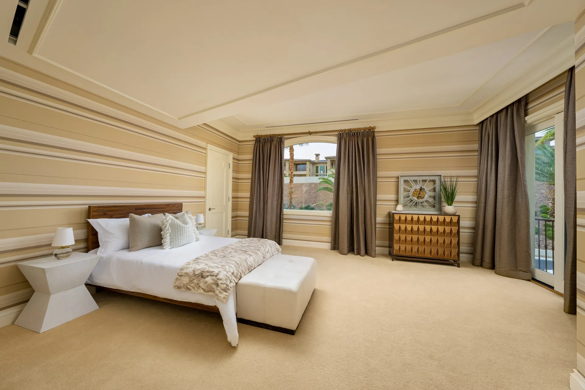 A bedroom with wide striped walls in neutral tones, featuring a wooden headboard, a white tufted bench at the foot of the bed, and a geometric chest. Brown drapes frame a large window overlooking the exterior.