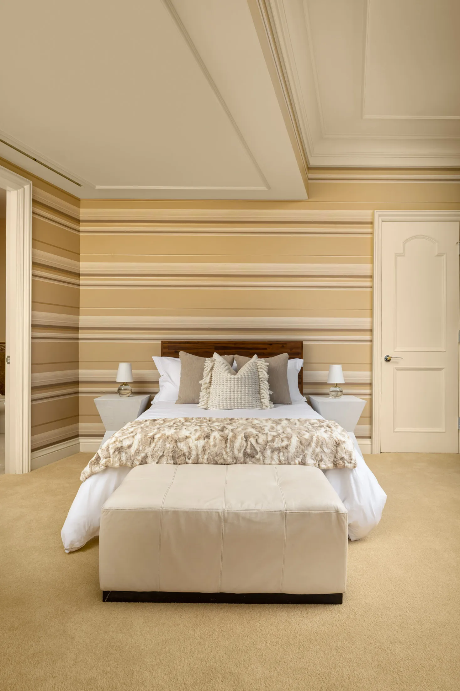 A bedroom with striped walls, a wooden headboard, neutral pillows, and a soft throw blanket. A padded bench sits at the foot of the bed.