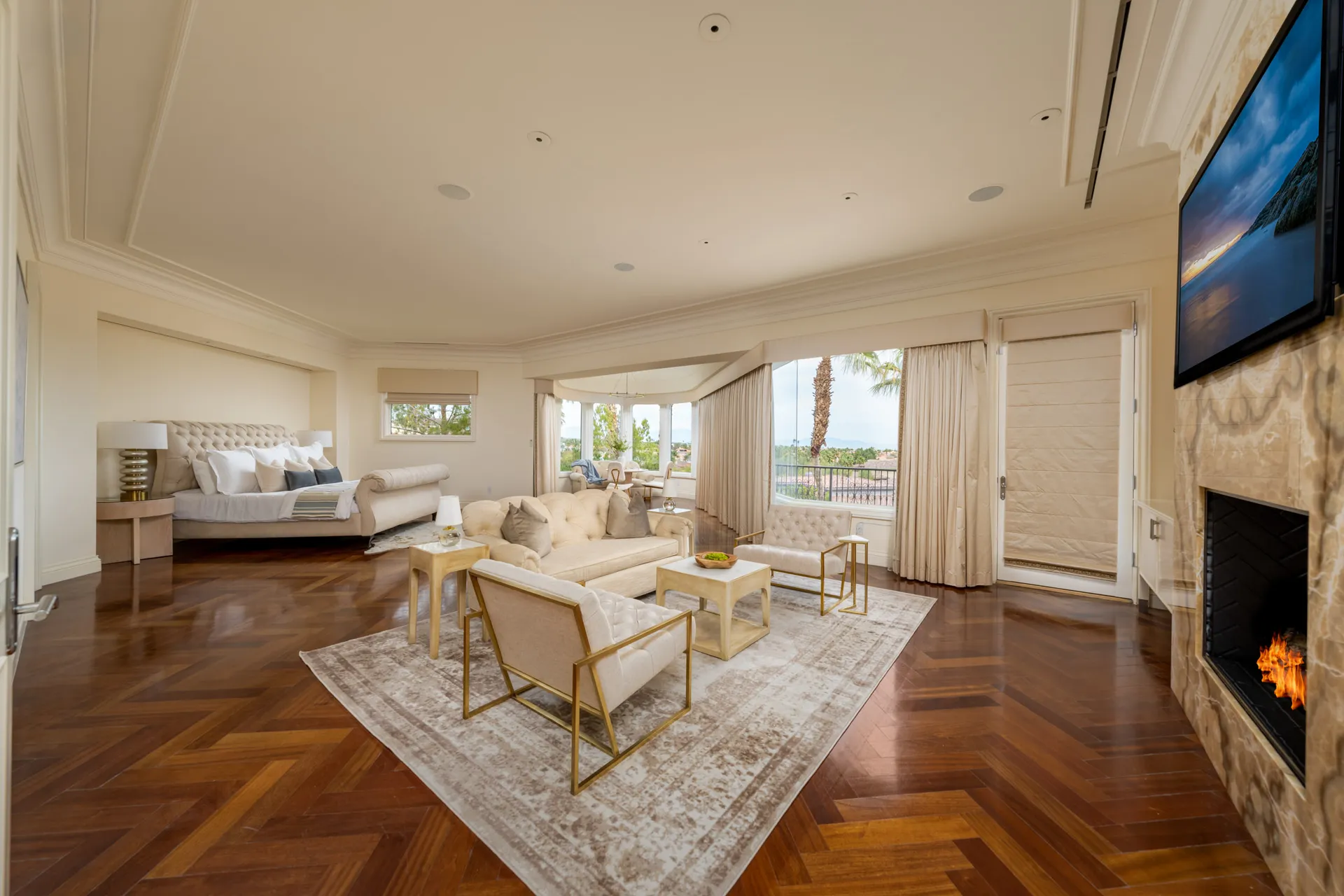 A luxurious bedroom with a tufted bed and a tufted seating area, large windows providing views of the surroundings, and a fireplace on the right. The floor features a rich herringbone pattern.