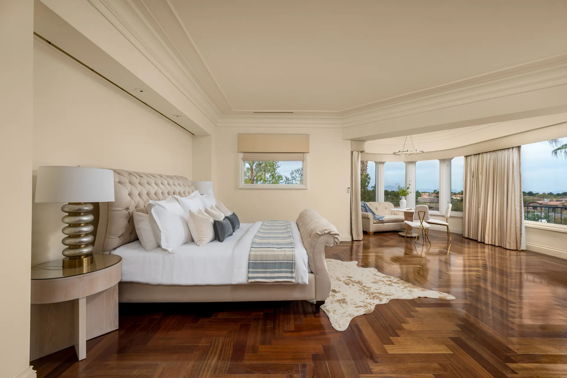A spacious bedroom featuring a tufted beige bed with layered pillows and herringbone wood floors. A curved window seating area provides sweeping views.