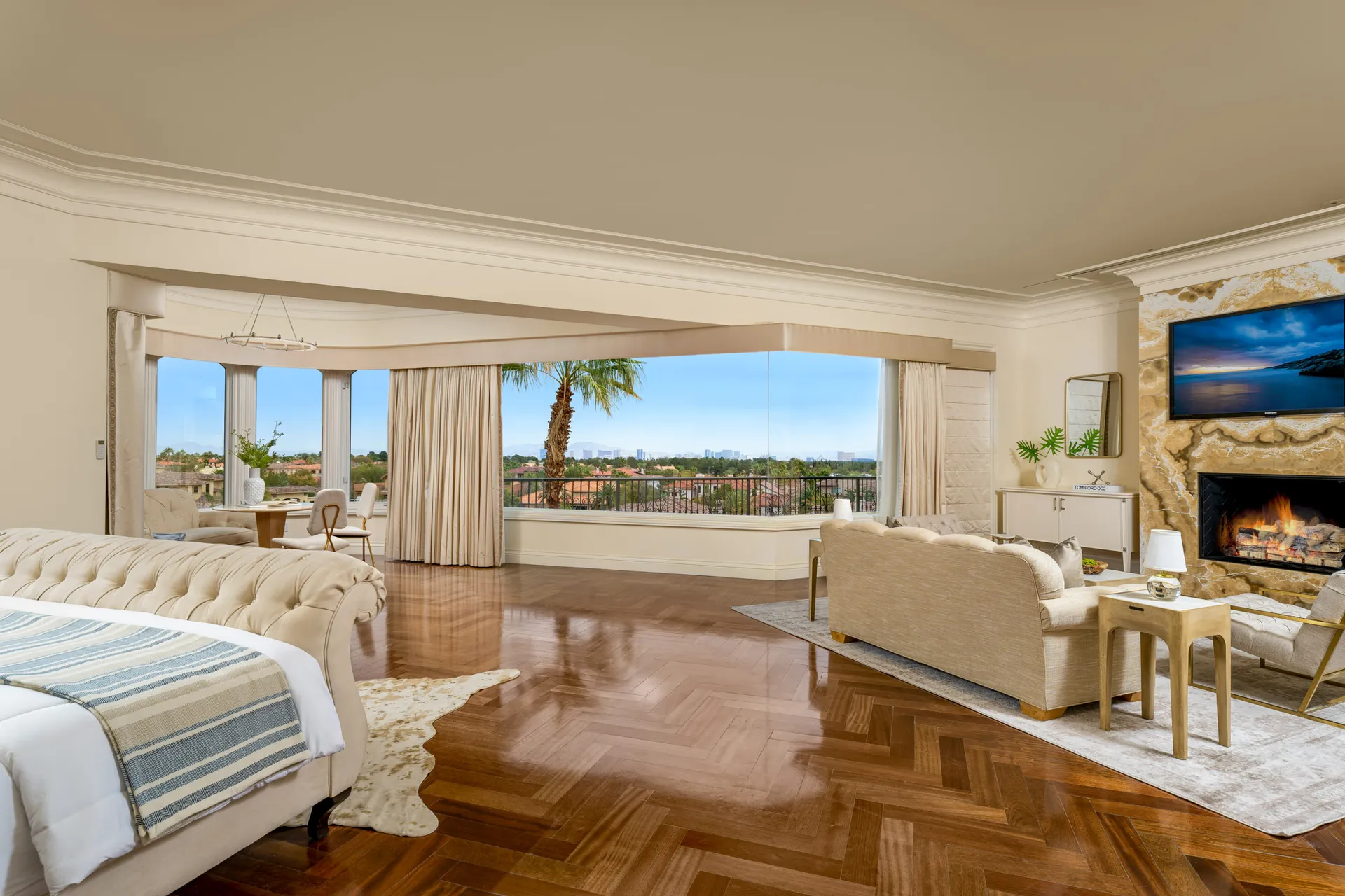 A luxurious bedroom suite featuring a tufted bed, large floor-to-ceiling windows overlooking a cityscape, and a seating area with a fireplace. Polished hardwood floors create a warm, elegant ambiance.