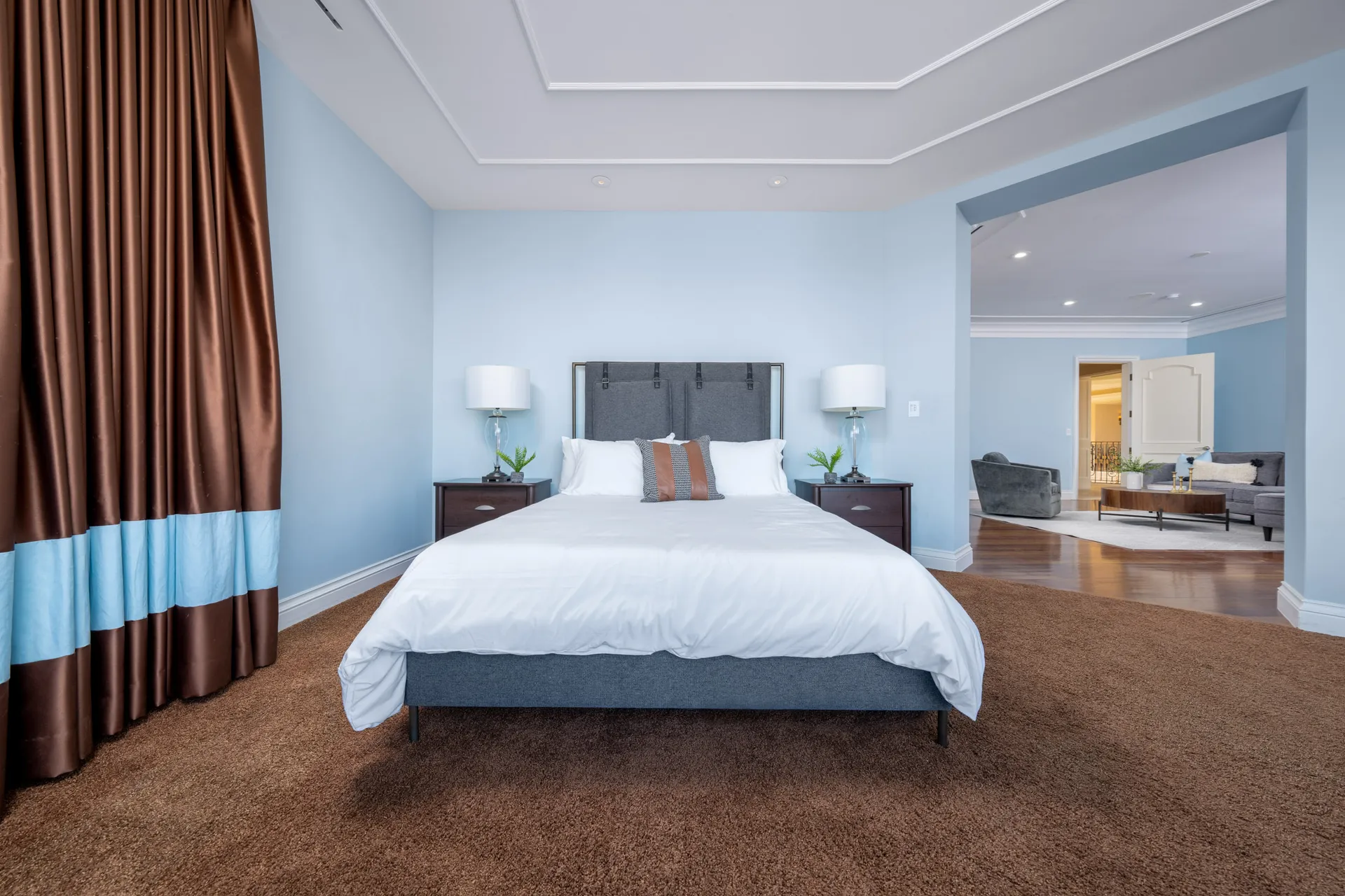 A bedroom with soft blue walls, a gray upholstered bed, and white linens. Brown-and-blue drapes frame one side, and an open archway leads to a seating area.
