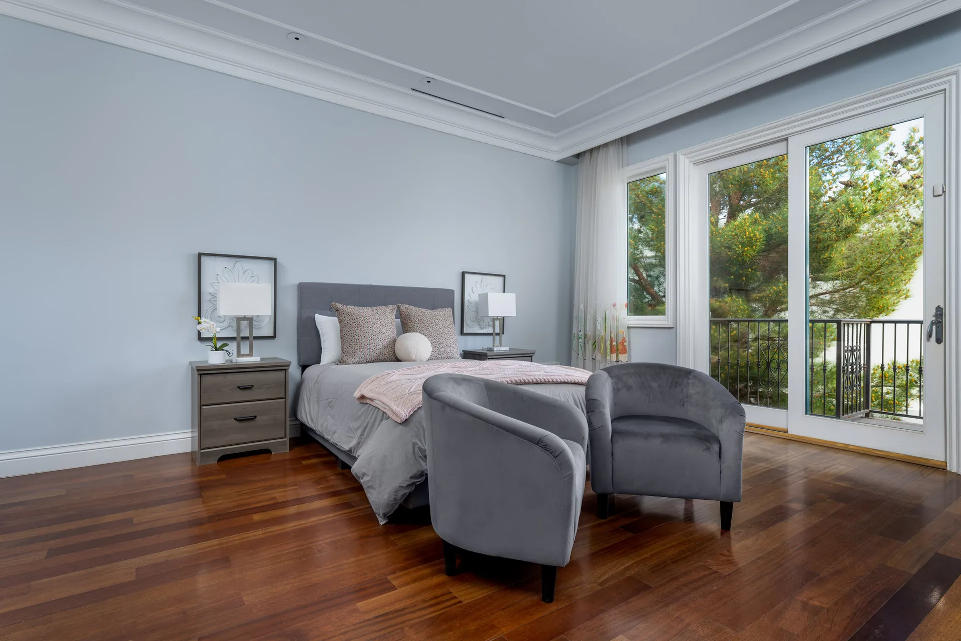 A softly lit bedroom featuring a gray upholstered bed with pink accents and two gray chairs beside large windows. Warm hardwood floors complete the cozy setting.