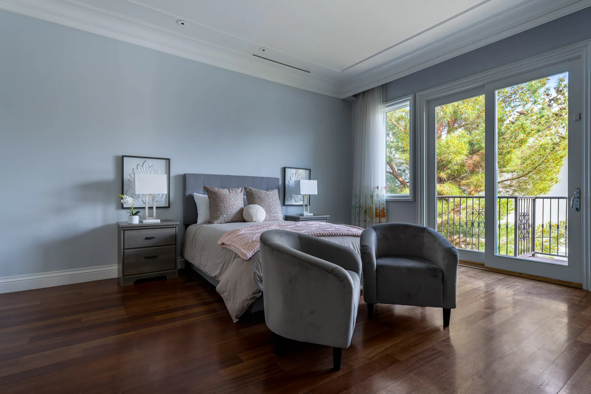 A cozy bedroom with a gray upholstered bed, pink accent pillows, and two gray chairs near large windows and a balcony. Rich wood floors add warmth.