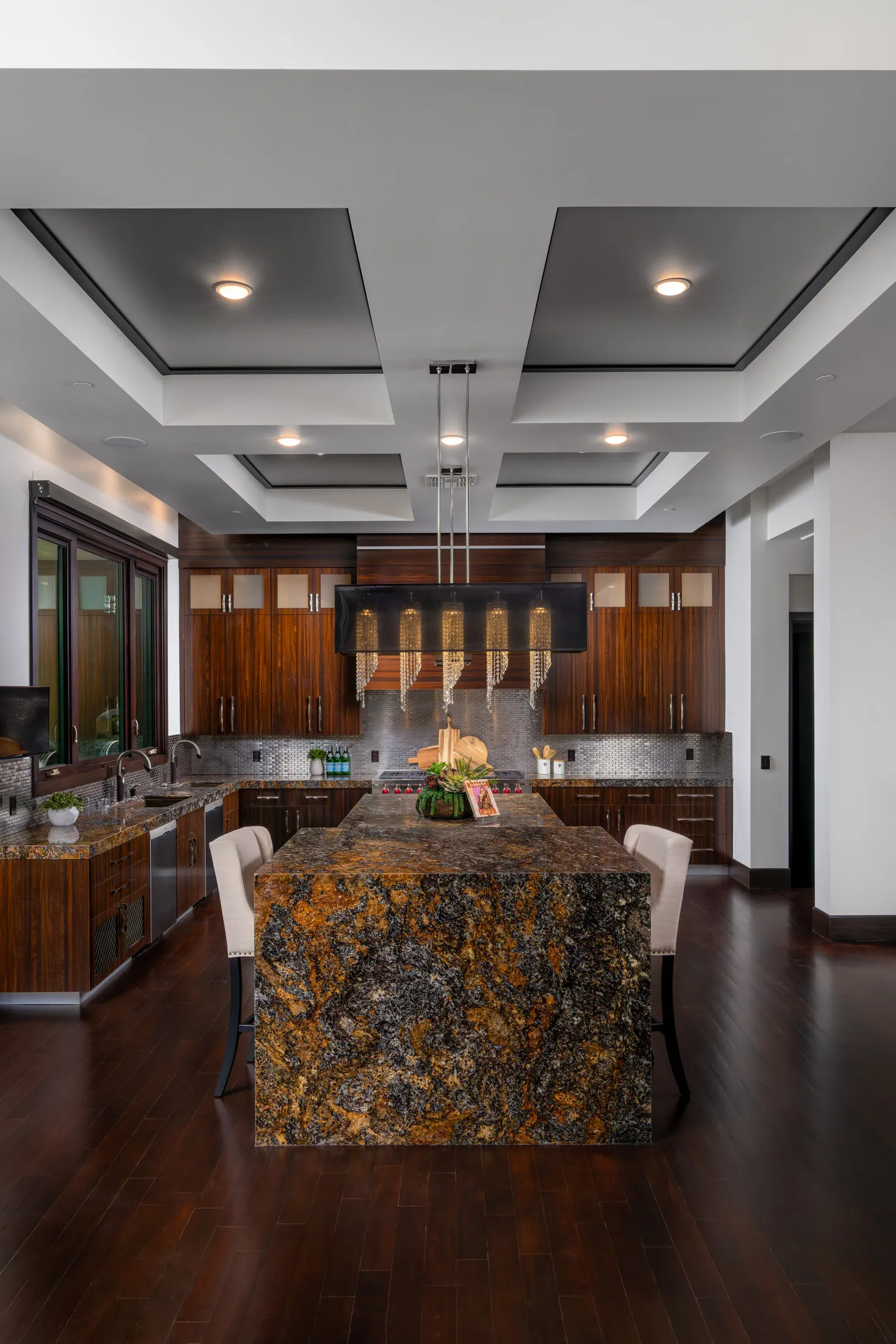 A modern kitchen featuring dark wood cabinets, a large granite island, and a statement chandelier under a coffered ceiling.