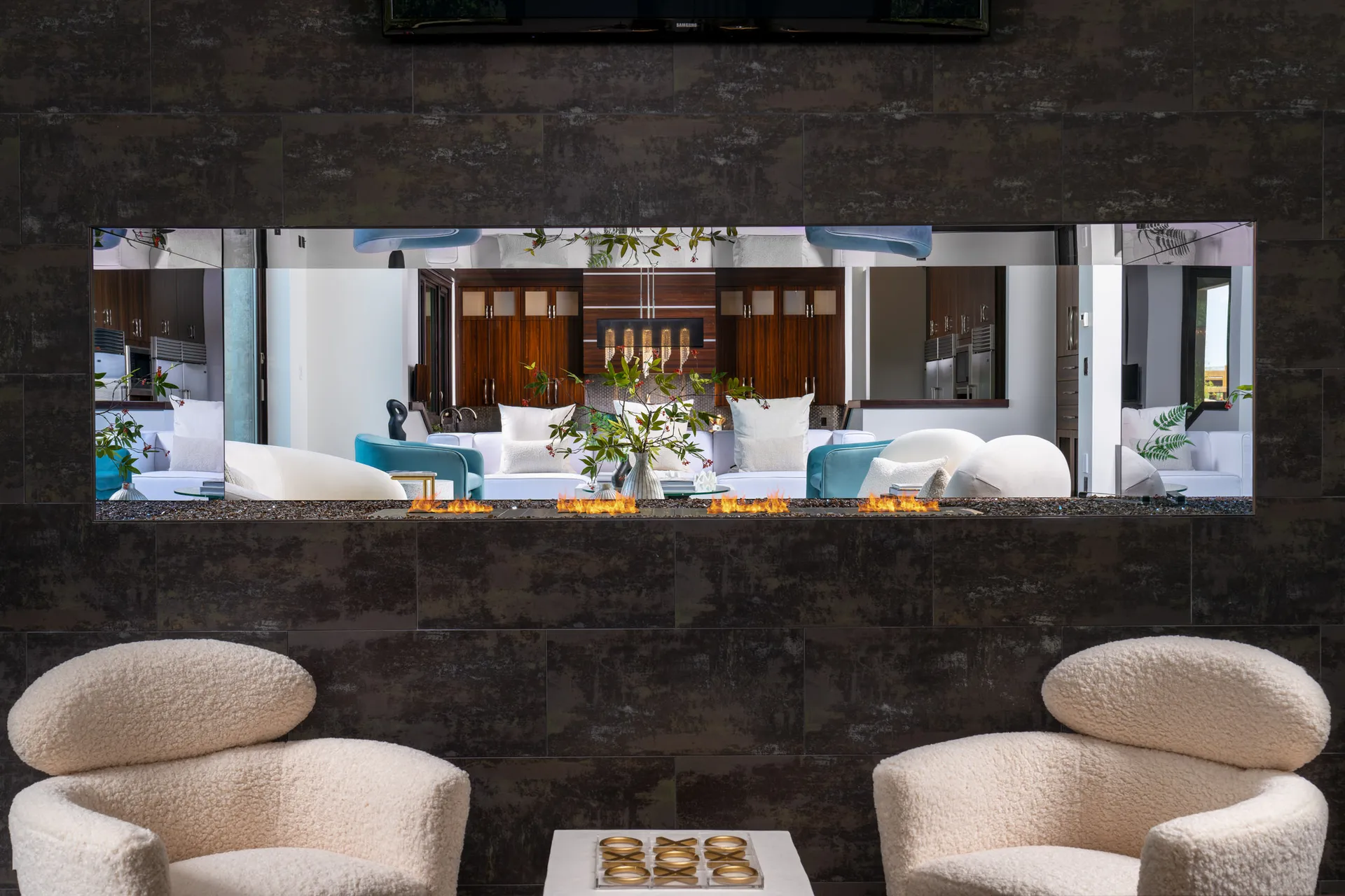 A dark tiled wall with a rectangular opening that reveals a fire feature, showcasing a modern seating area with teal and white furnishings beyond