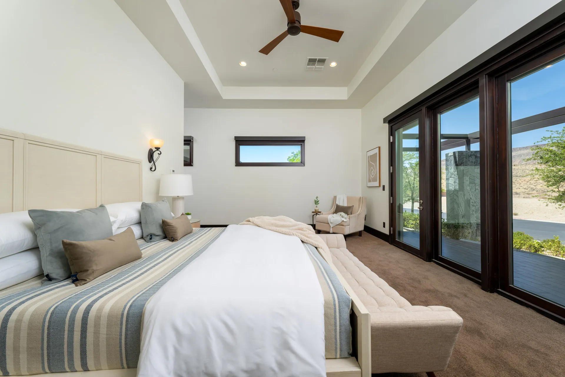 A serene bedroom with a neutral bed, corner seating, and large windows opening to an outdoor view.