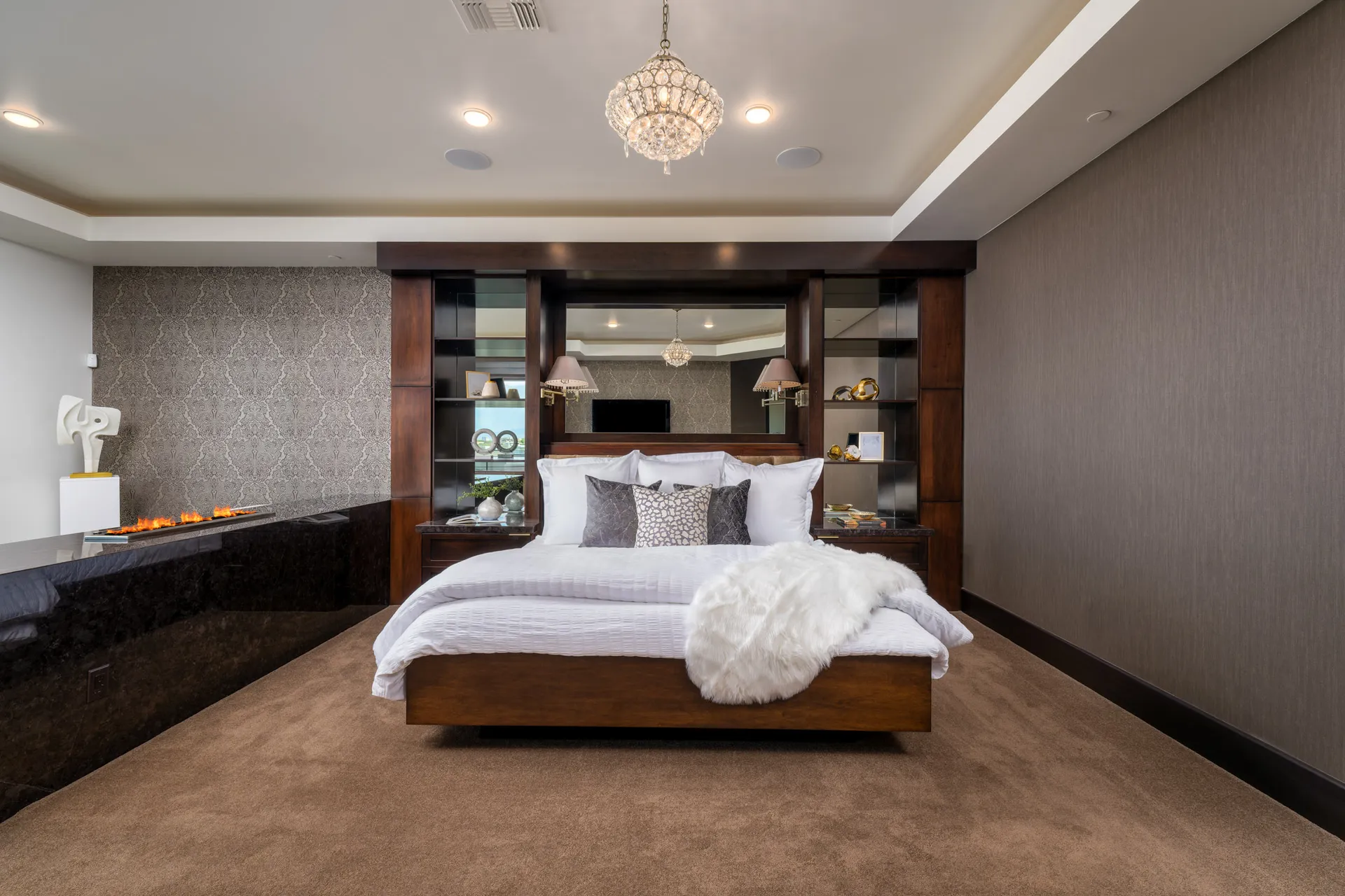 A warm bedroom with a wooden bed frame, built-in shelving behind the headboard, and a sleek fireplace under a crystal chandelier.