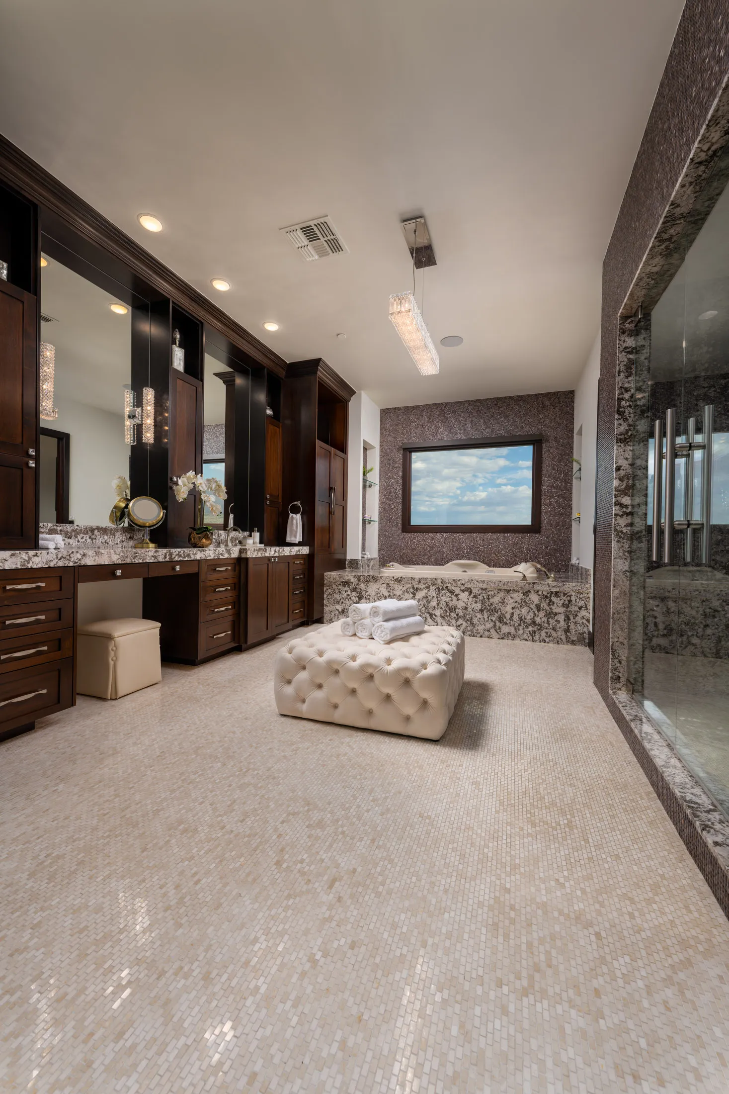 Luxurious bathroom with large vanity, marble countertops, tufted ottoman, and walk-in shower.