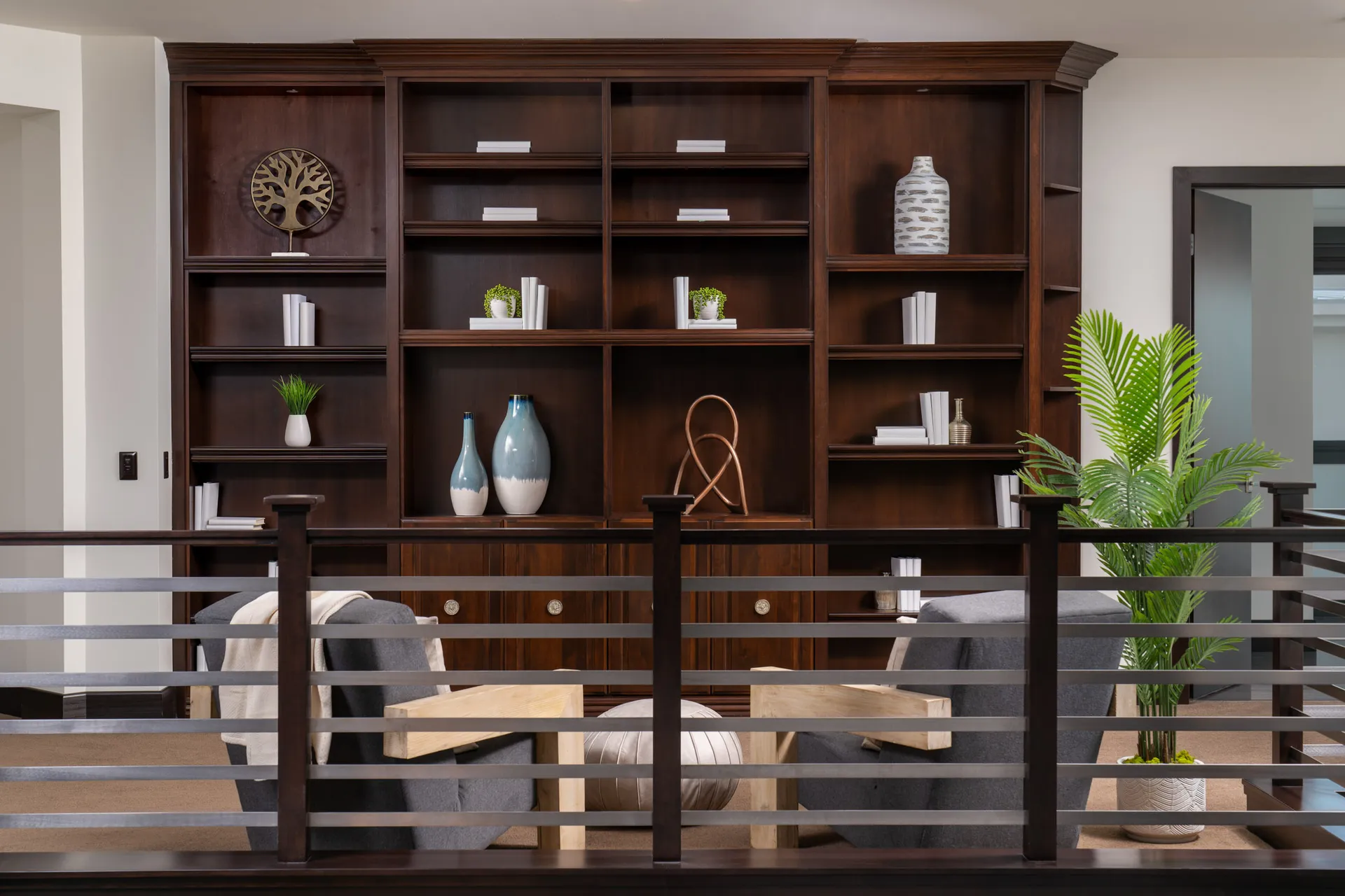Loft area with wooden bookshelf, gray armchairs, round ottoman, and wooden railing.