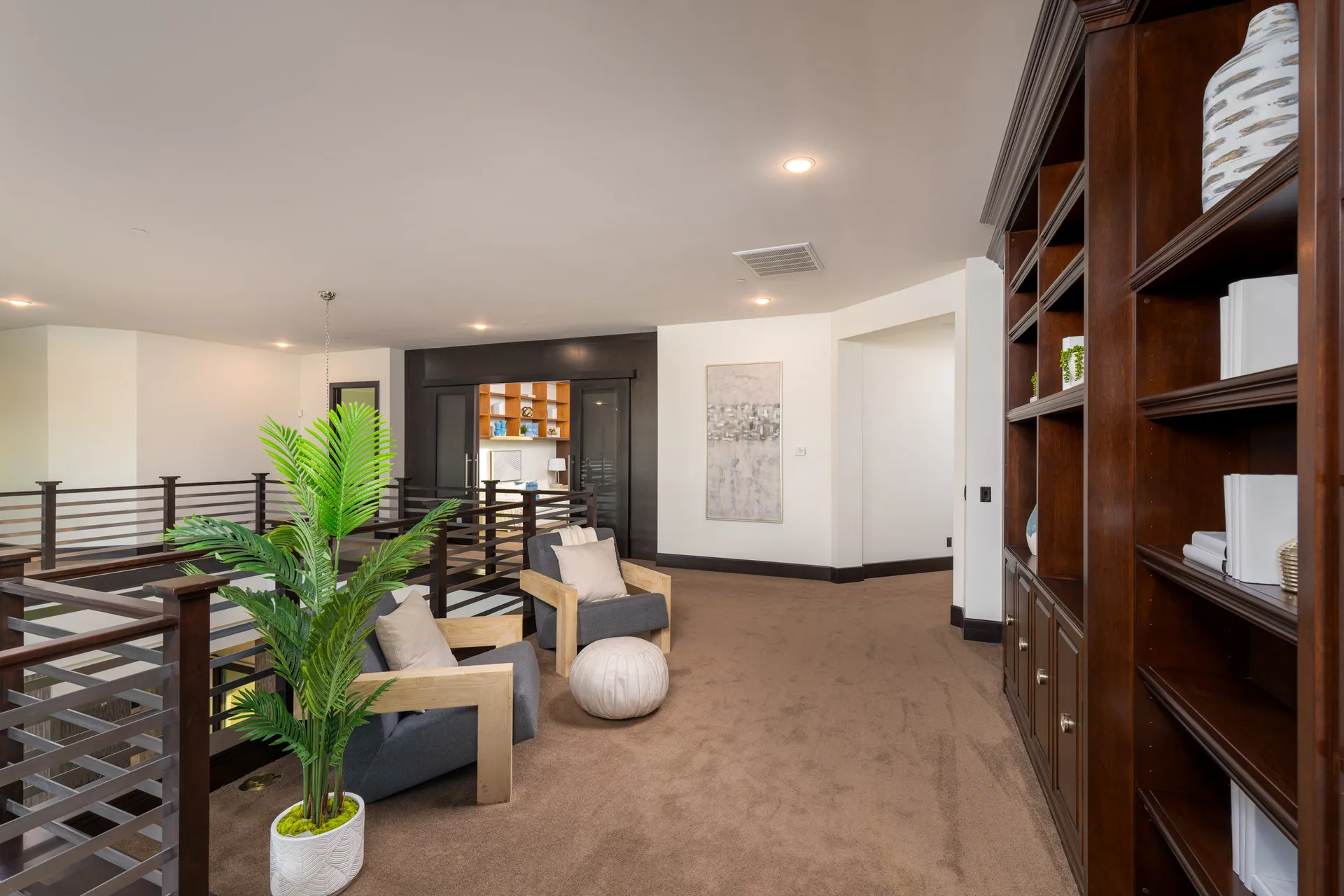 Loft area with gray armchairs, round ottoman, wooden railing, and built-in wooden bookshelf.