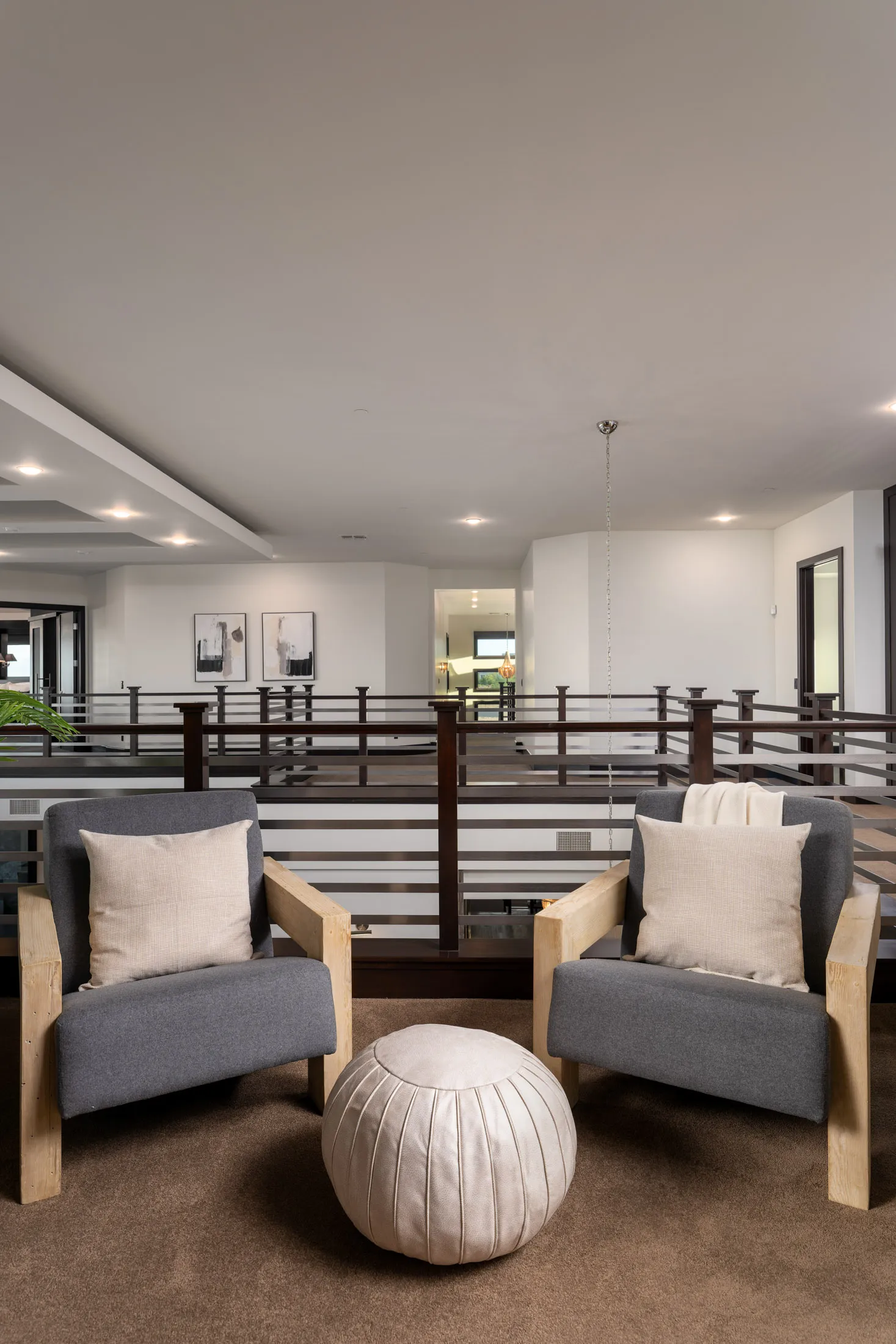 Modern living room with gray armchairs, round ottoman, and wooden railing.
