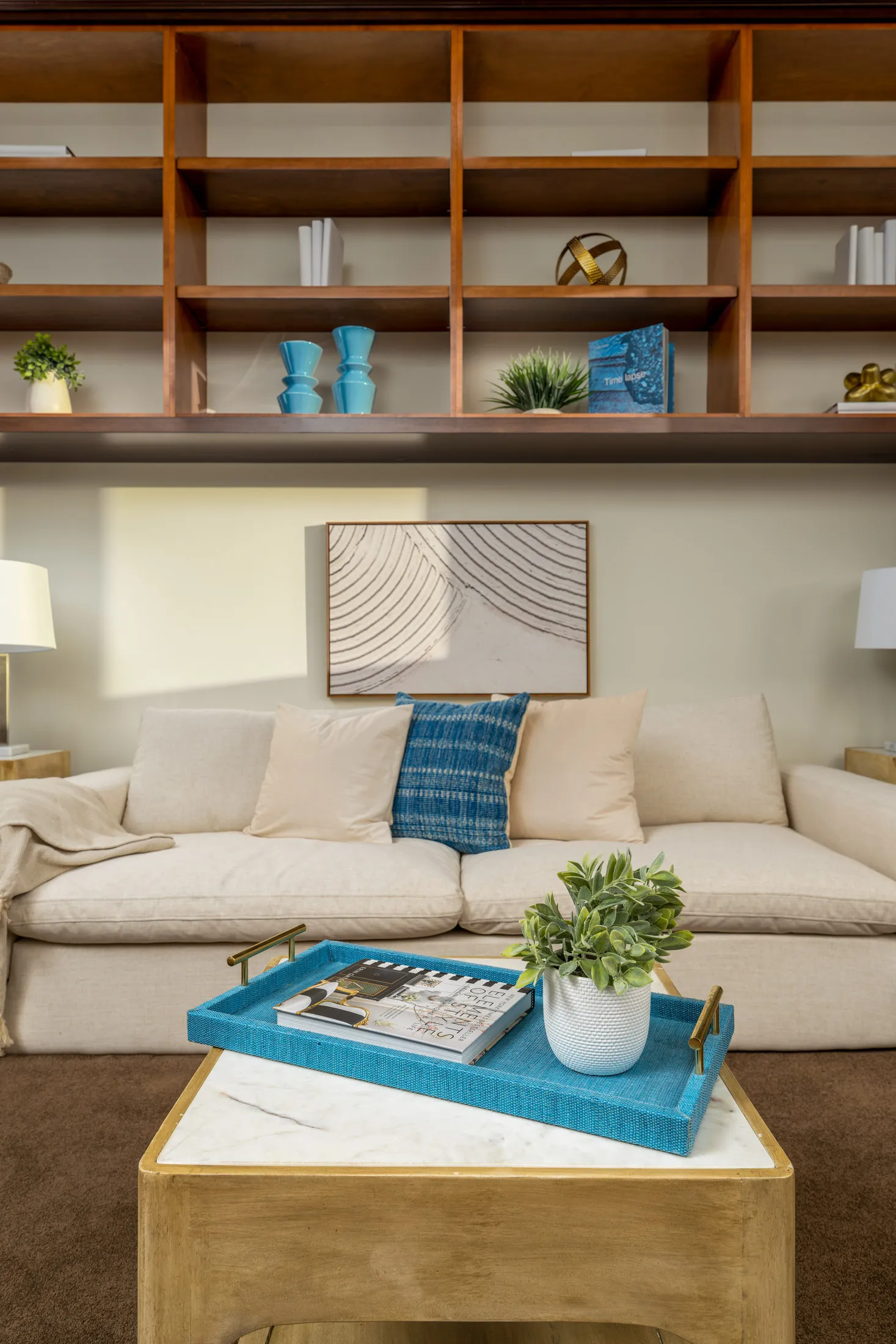 Cozy living room with beige sofa, blue throw blanket, marble coffee table, and wooden bookshelf.