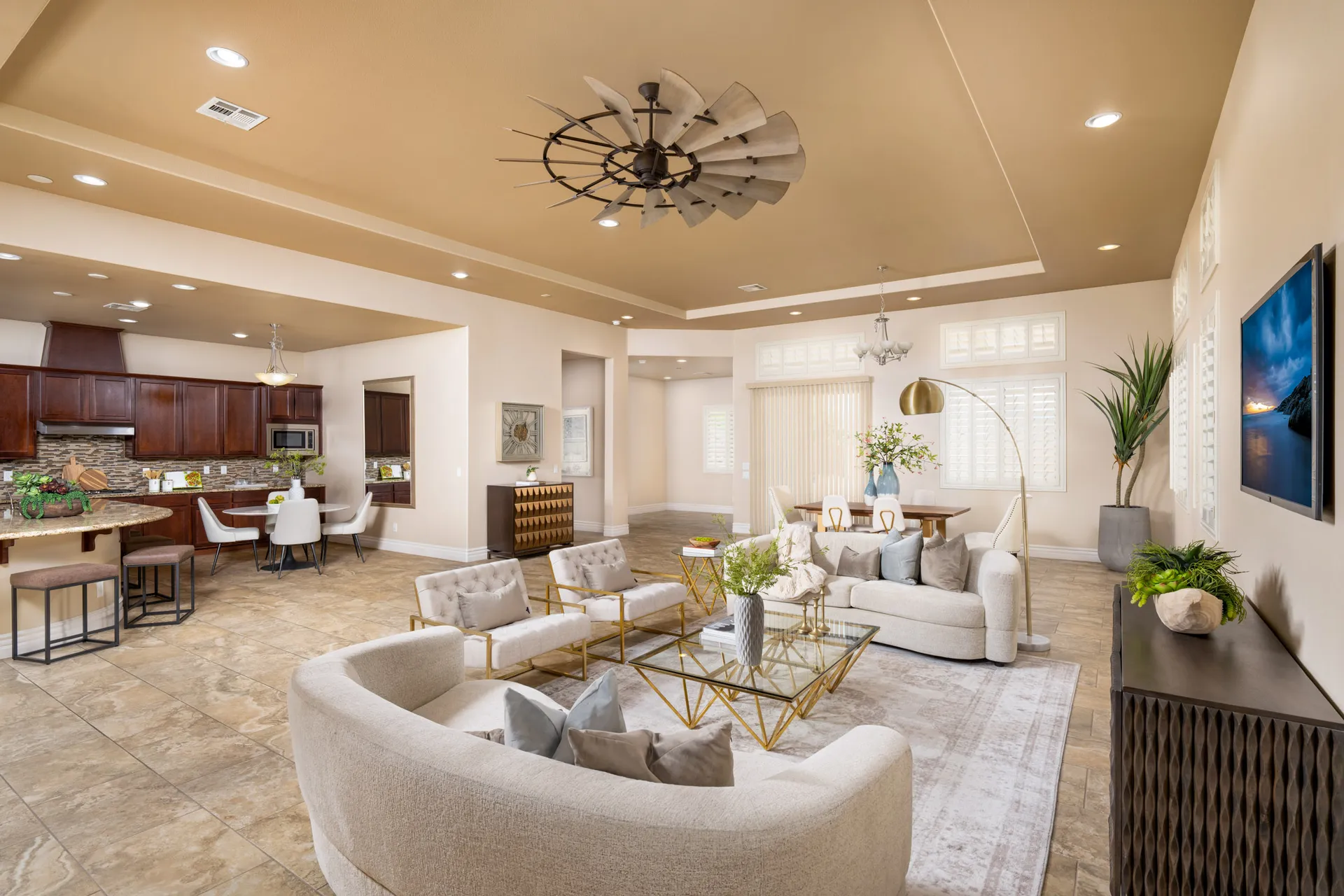 Cozy Home Staging An open-concept area with a curved sofa, gold-toned chairs, and a wooden dining table. A large windmill-style ceiling fan hangs above.