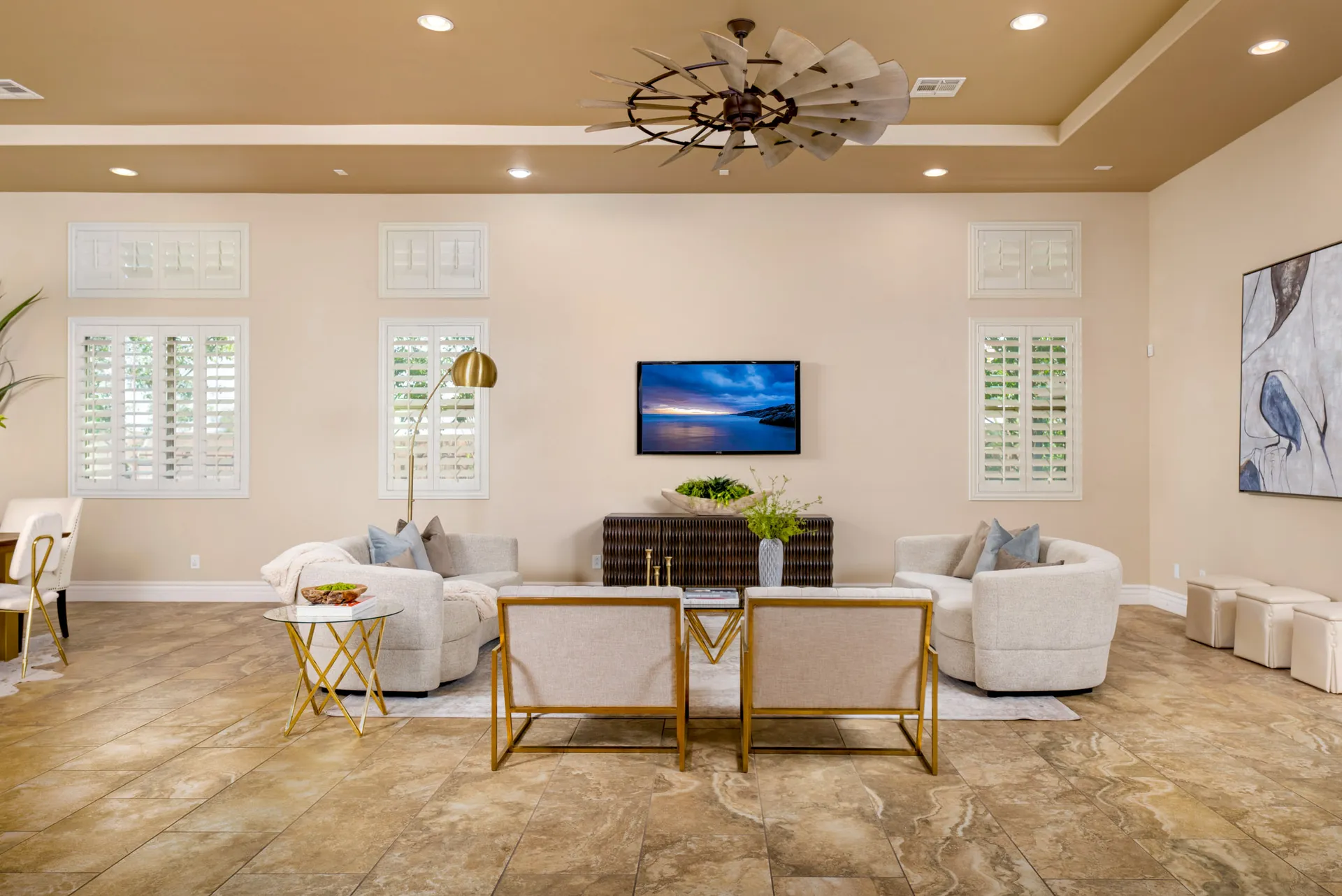 A light-filled living area with a curved sofa, gold-framed chairs, and a windmill-style ceiling fan, centered around a wall-mounted TV above a textured console.