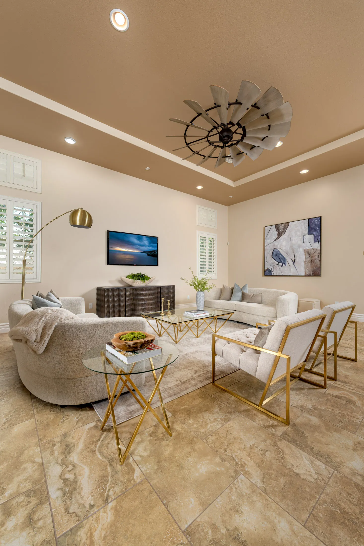 A spacious living area featuring a curved neutral sofa, gold-framed chairs and tables, and a large windmill-style ceiling fan.