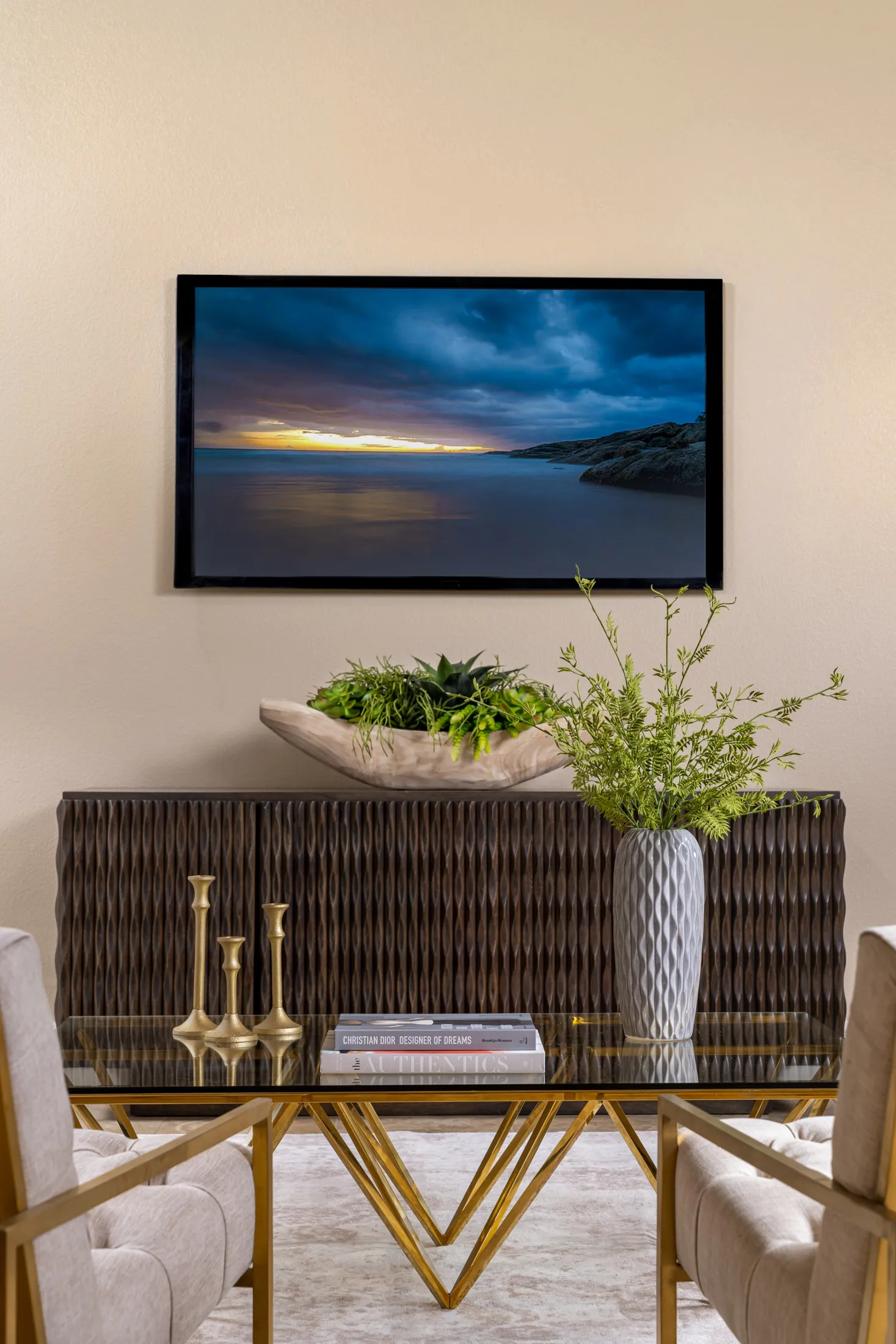 A carved wood console topped with greenery sits below a wall-mounted TV showing a coastal sunset, with a glass coffee table and gold accents in front.