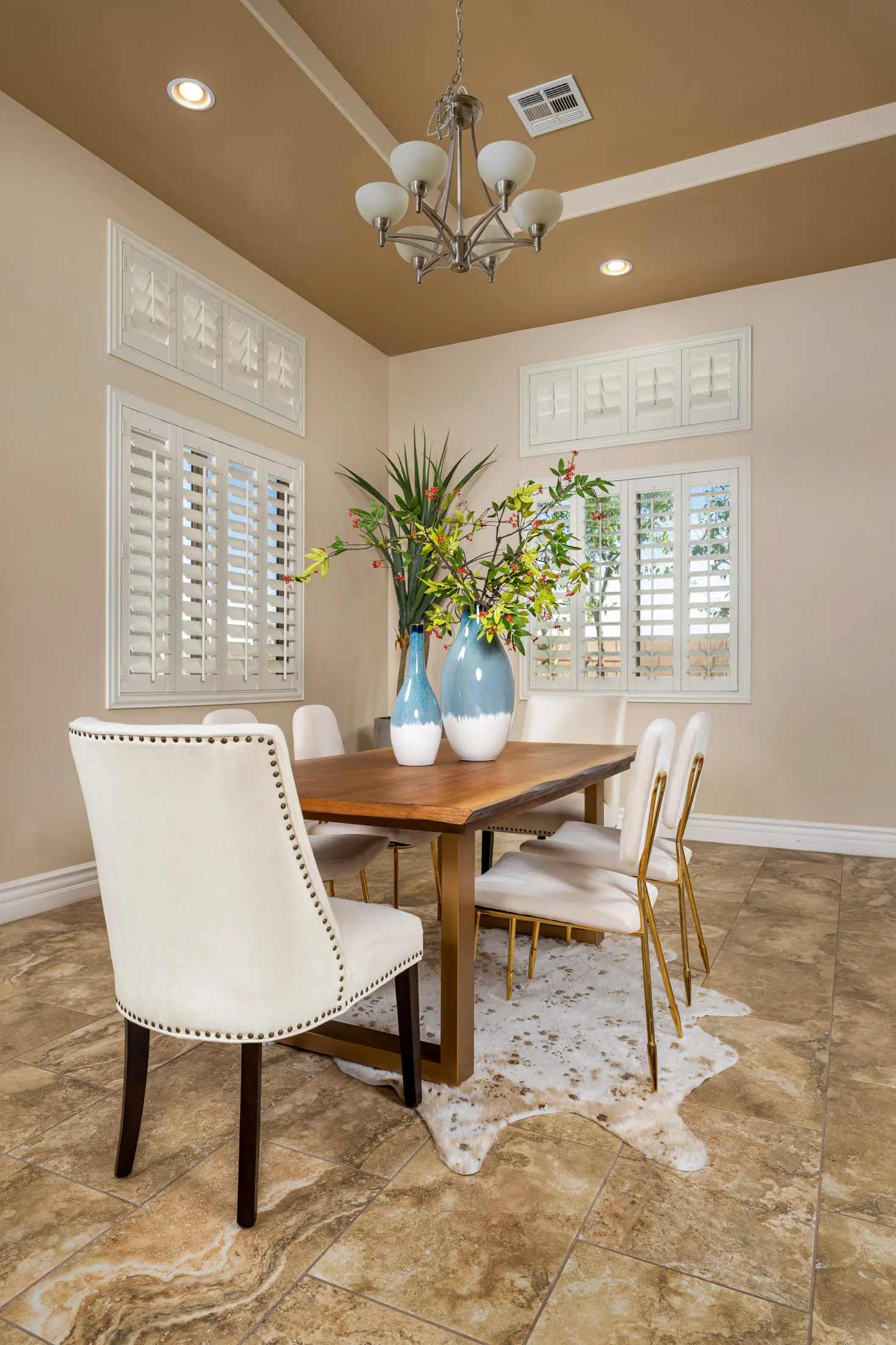 A warm dining room with a wood table, white chairs, tall blue vases of greenery on a hide rug, and a modern chandelier overhead.
