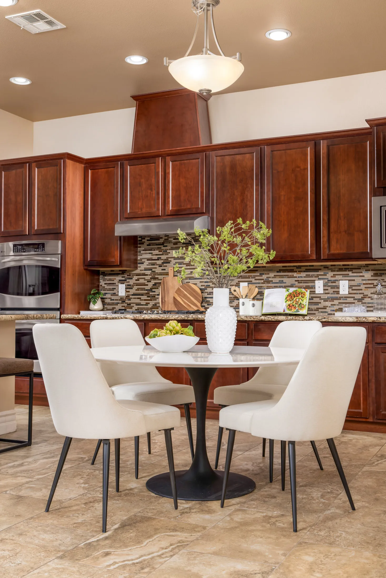 A modern dining area with a round white table, upholstered chairs, and a mosaic backsplash in a wood-cabinet kitchen.