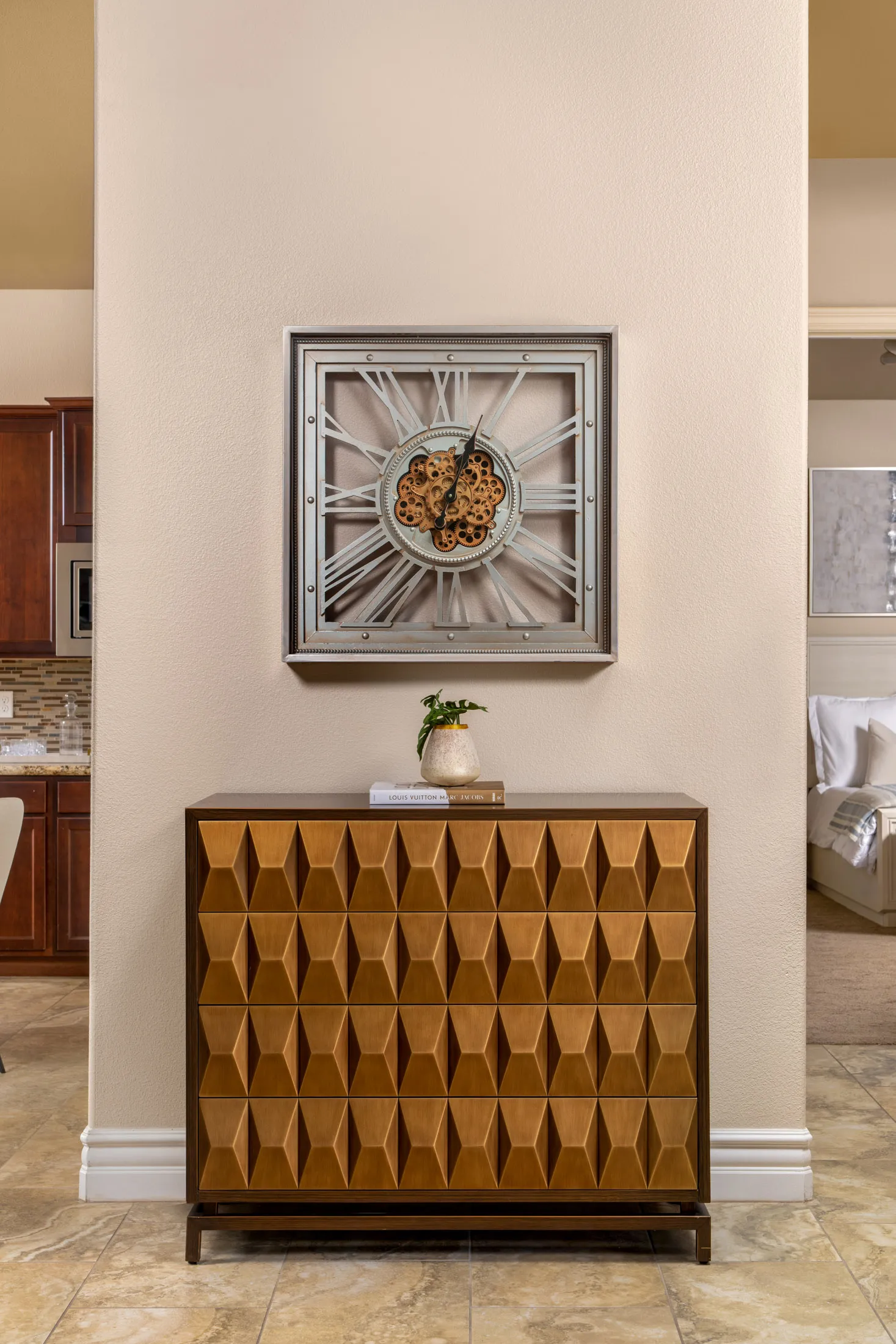 A wooden chest with a three-dimensional pattern, topped by books and a small plant, beneath a large metal clock with exposed gears.