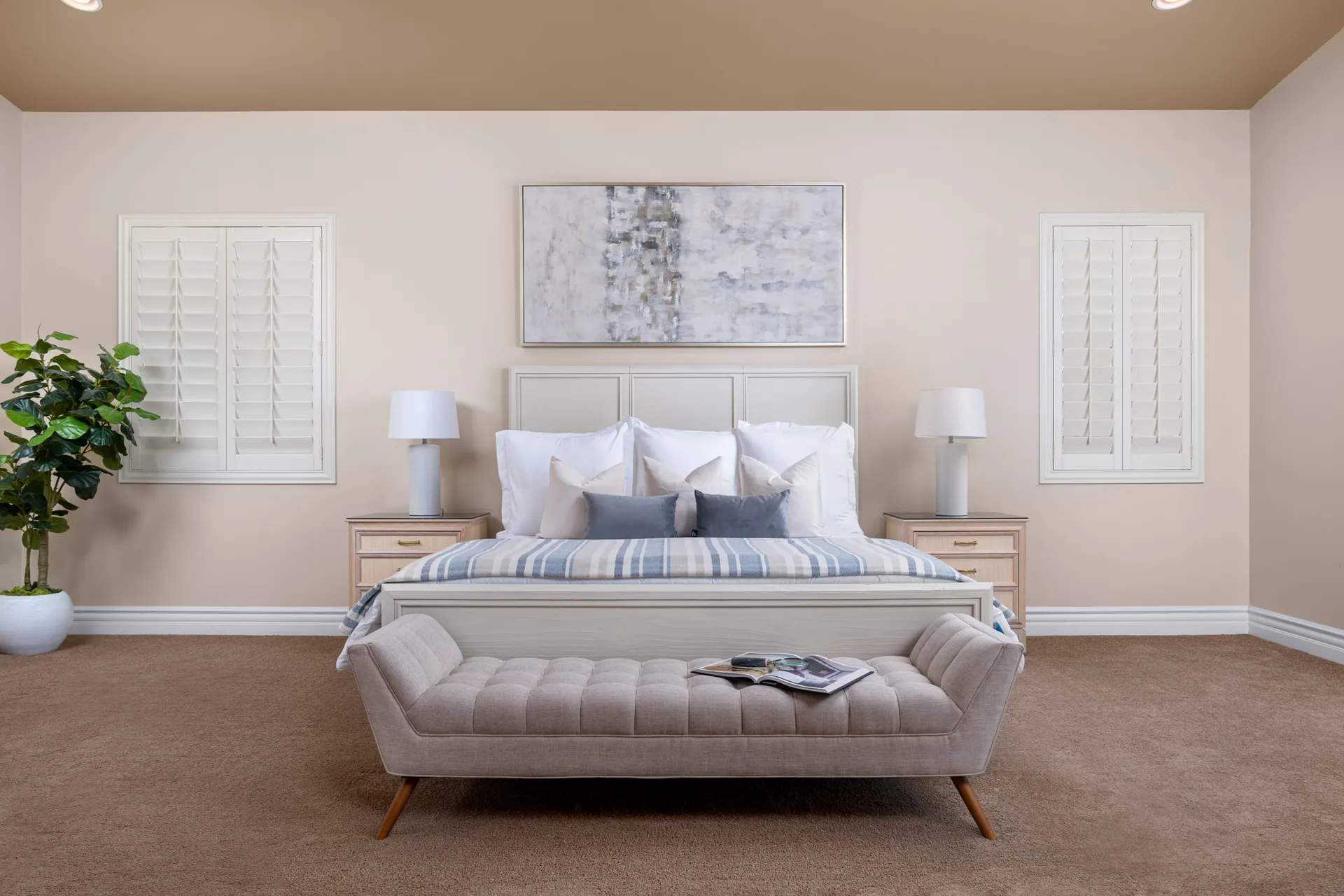 A neutral bedroom with a tufted bench, striped blanket, and side tables with lamps, set under a large abstract painting and framed by shuttered windows.
