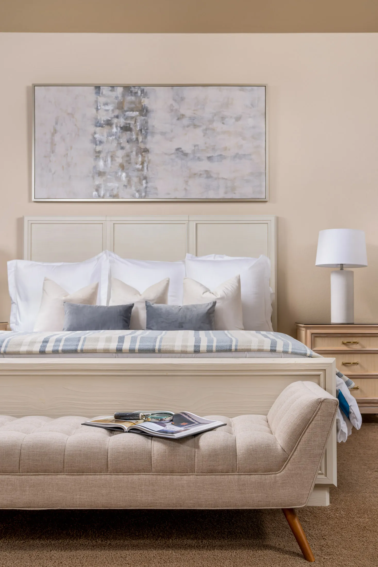 A light-toned bed with layered pillows, a striped blanket, and a tufted bench at the foot under a large abstract painting.