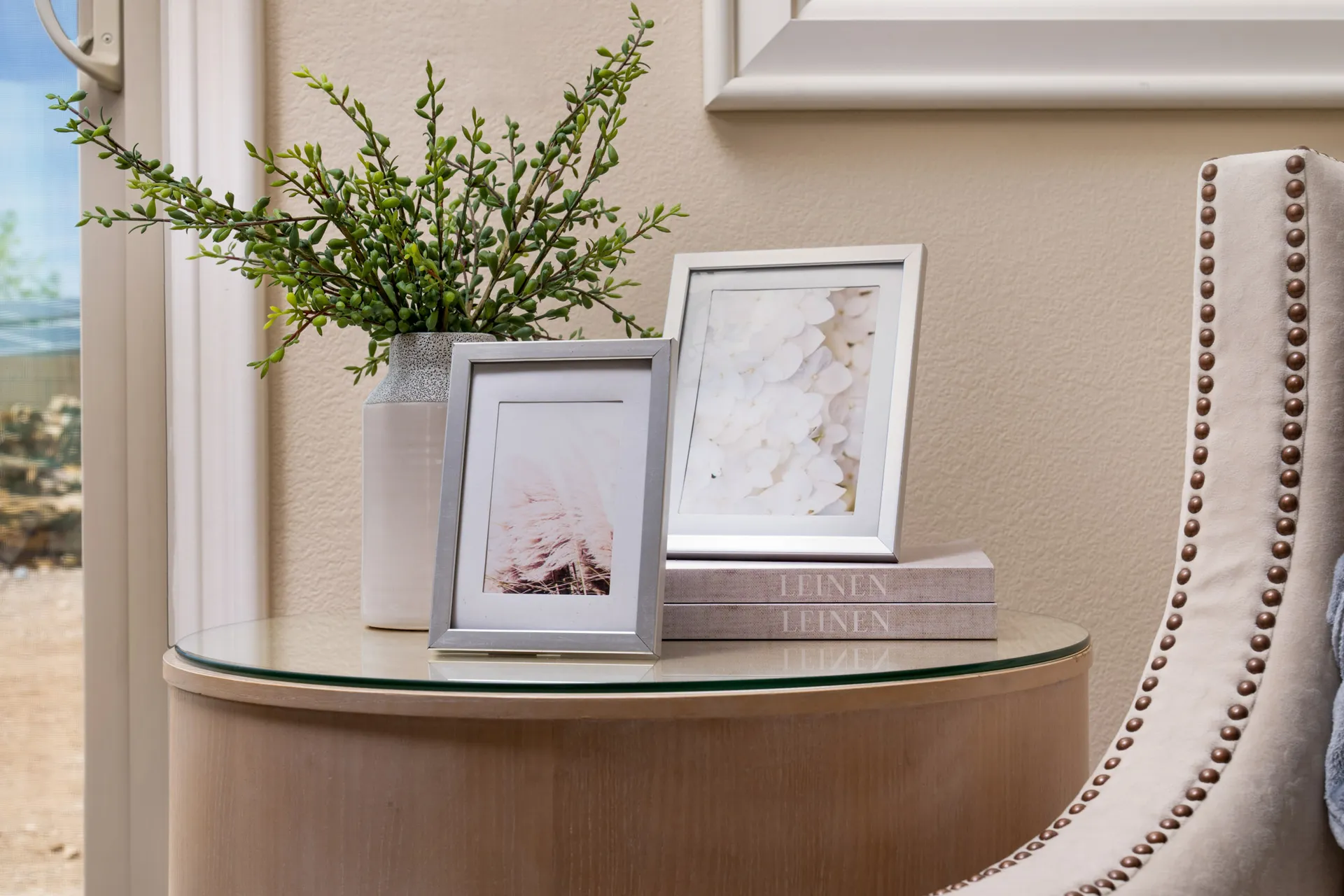 A round side table with a glass top holding silver-framed photos, a vase of greenery, and stacked books next to a studded neutral chair.