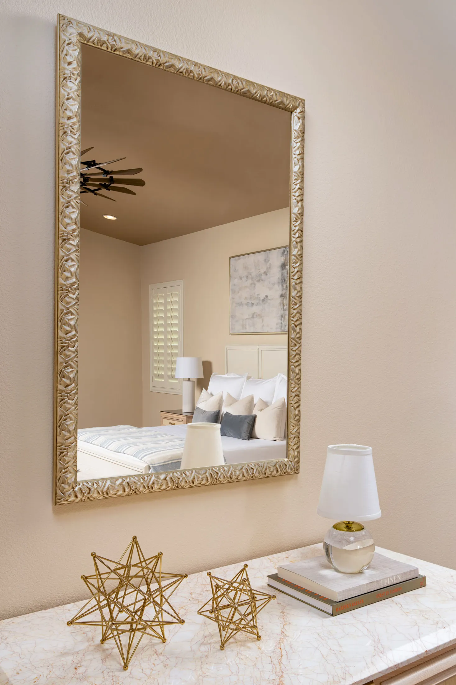 A gold-framed mirror reflecting a bed with neutral pillows, above a console table with metallic star sculptures and a small lamp.
