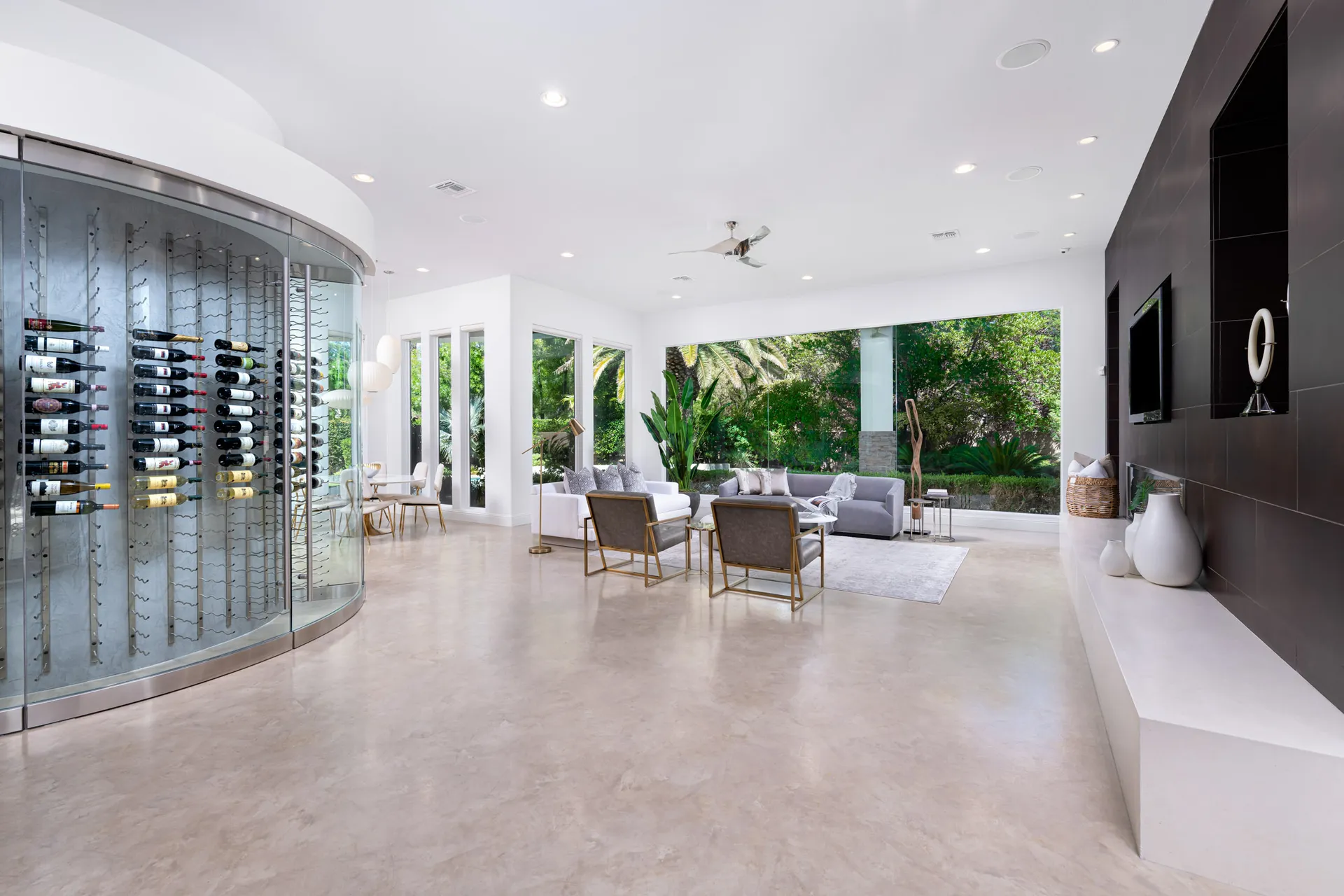 A fully staged open-concept living room featuring elegant seating, a statement wine display, and floor-to-ceiling windows with lush outdoor views.