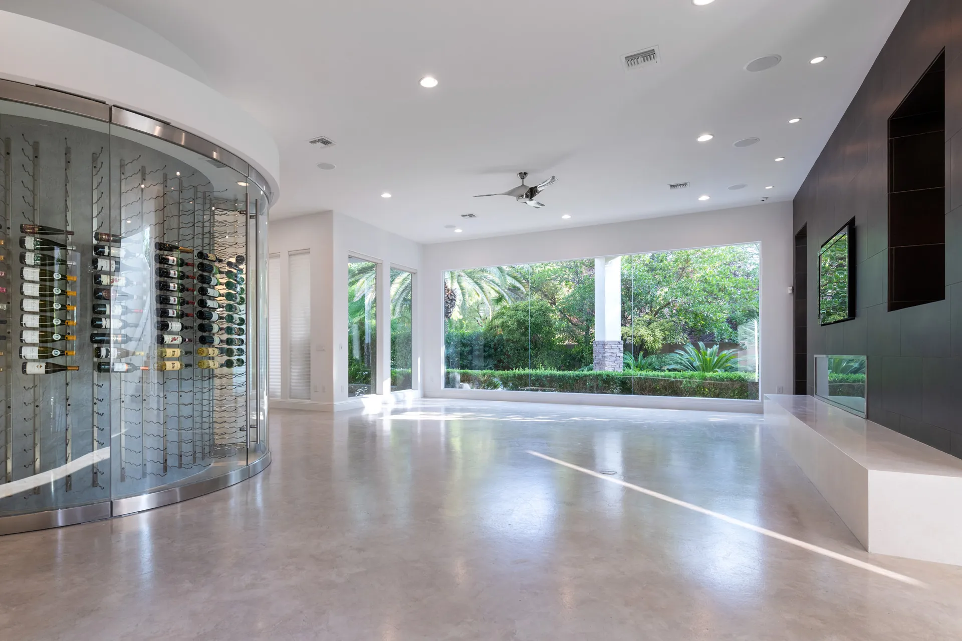 An empty modern living room featuring a glass wine display, a dark accent wall with a built-in fireplace, and floor-to-ceiling windows overlooking greenery.