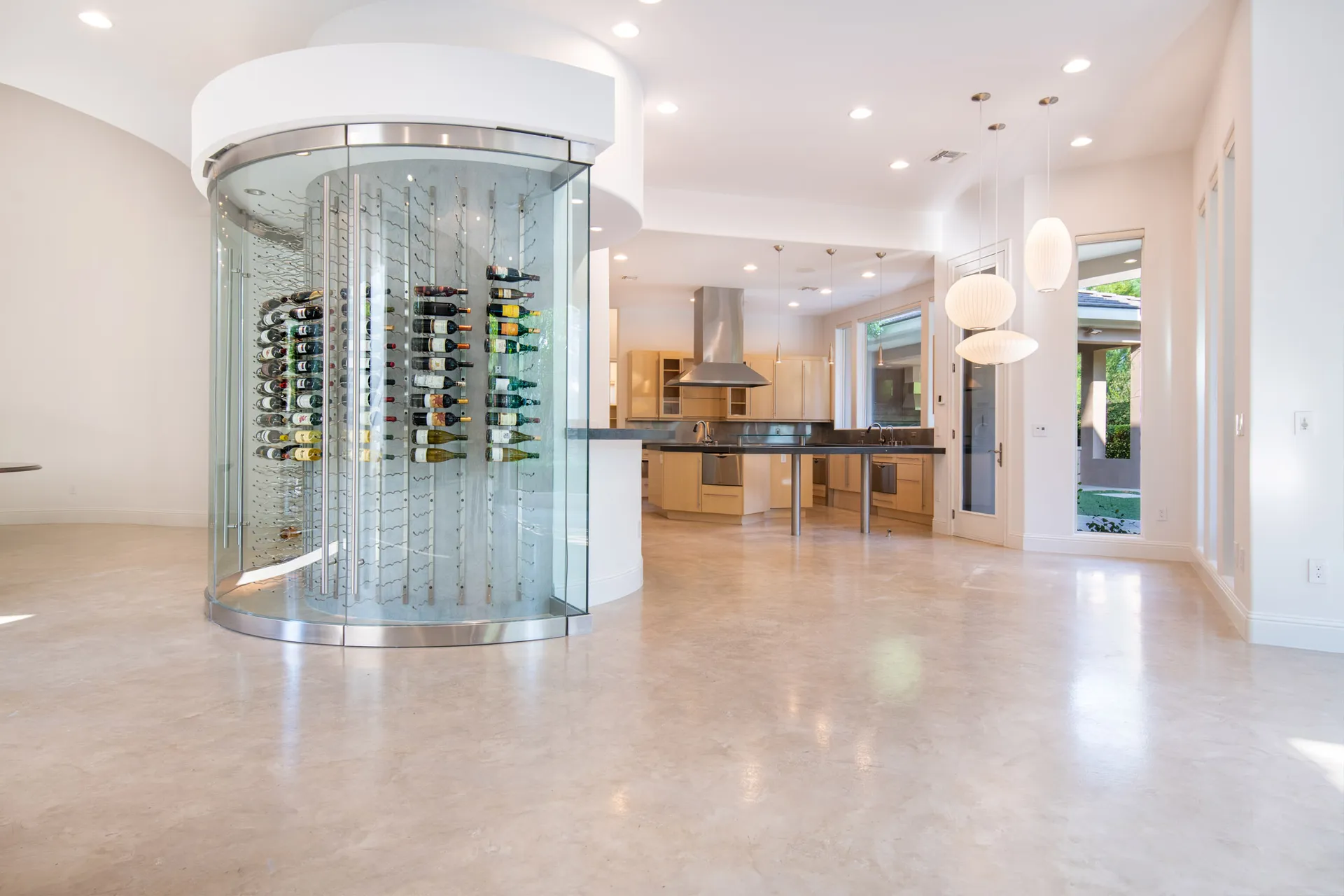 An empty living space featuring a modern glass wine display, open kitchen, and large windows bringing in natural light.