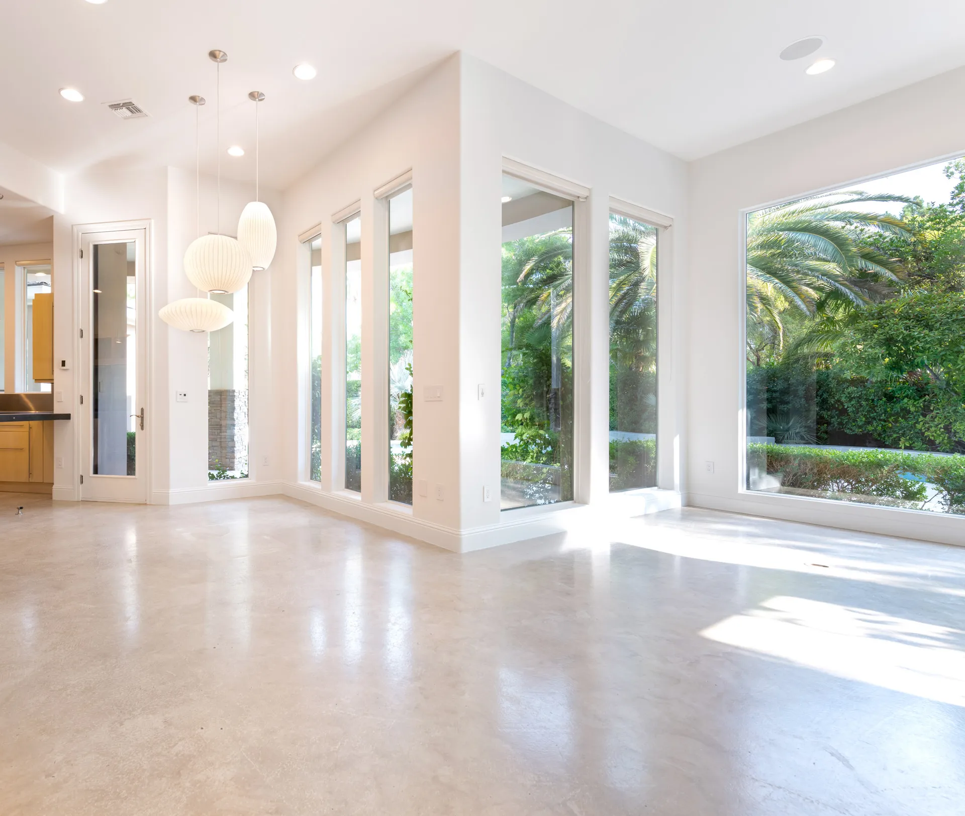 An empty living room with floor-to-ceiling windows, white walls, and natural light overlooking lush greenery.