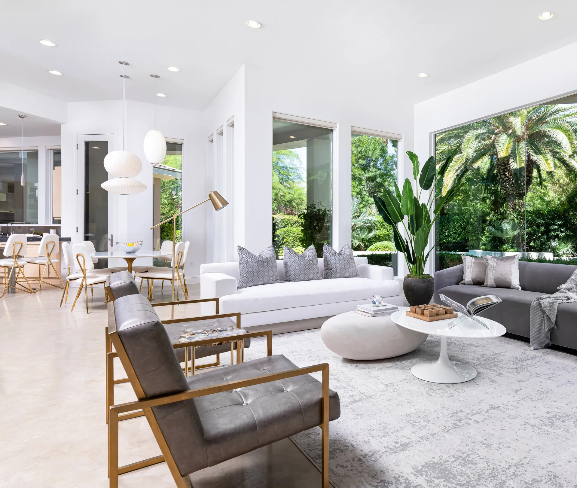 A modern living room with floor-to-ceiling windows, plush seating, gold-accented chairs, and lush greenery visible through large glass panels.