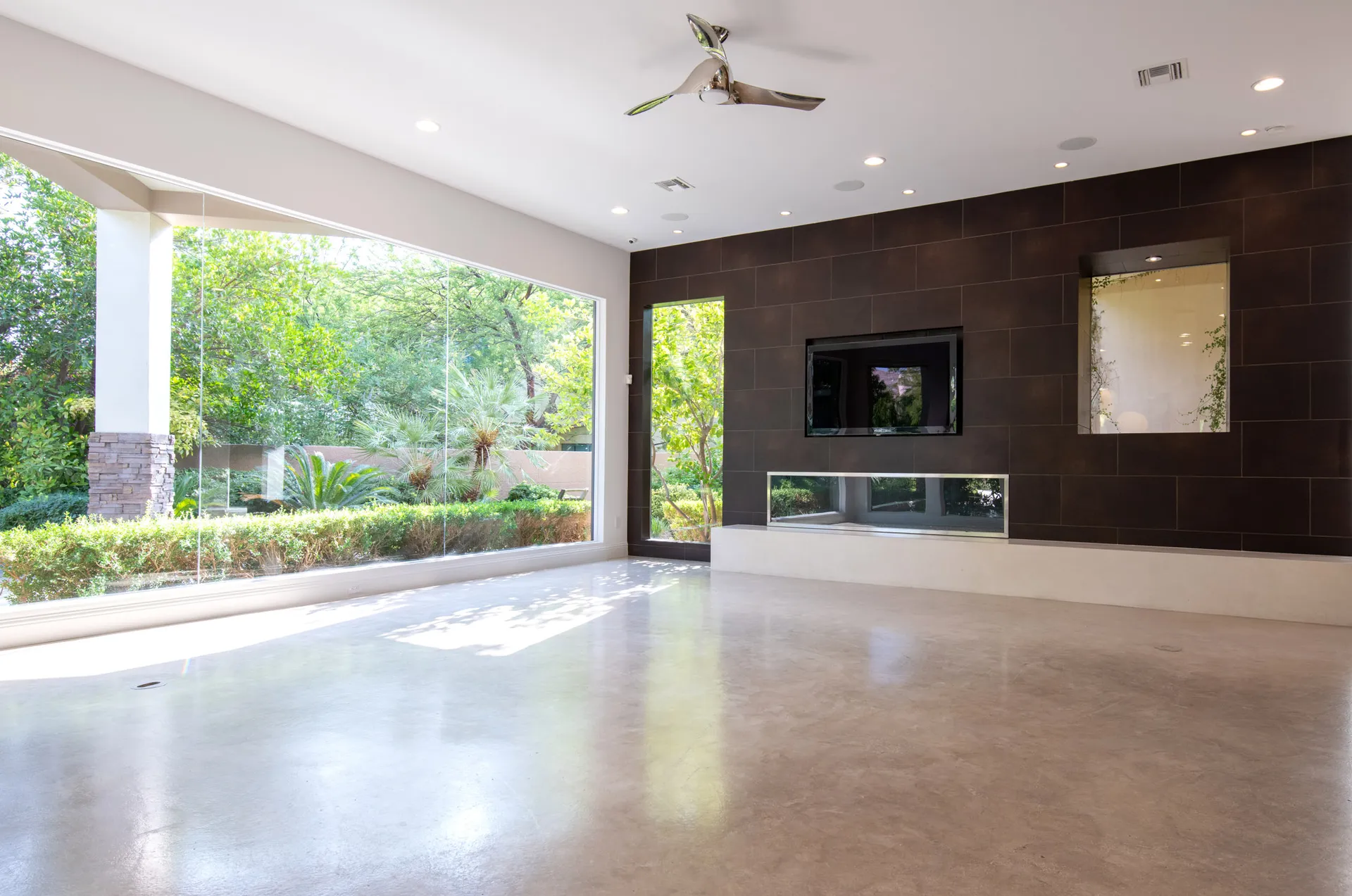 An empty modern living room with floor-to-ceiling windows, a dark accent wall, and a built-in fireplace overlooking lush greenery.