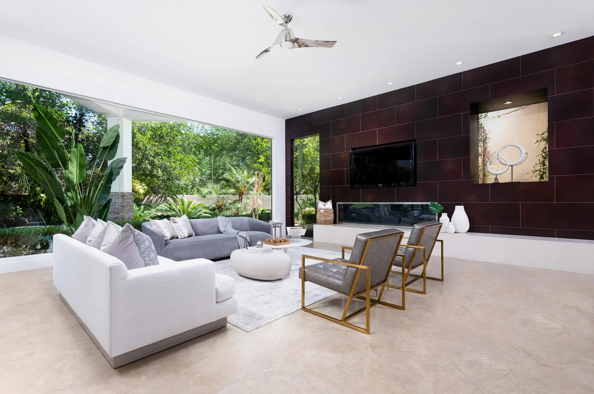 A modern living room with floor-to-ceiling windows, plush seating, a sleek fireplace, and a dark accent wall overlooking lush greenery.