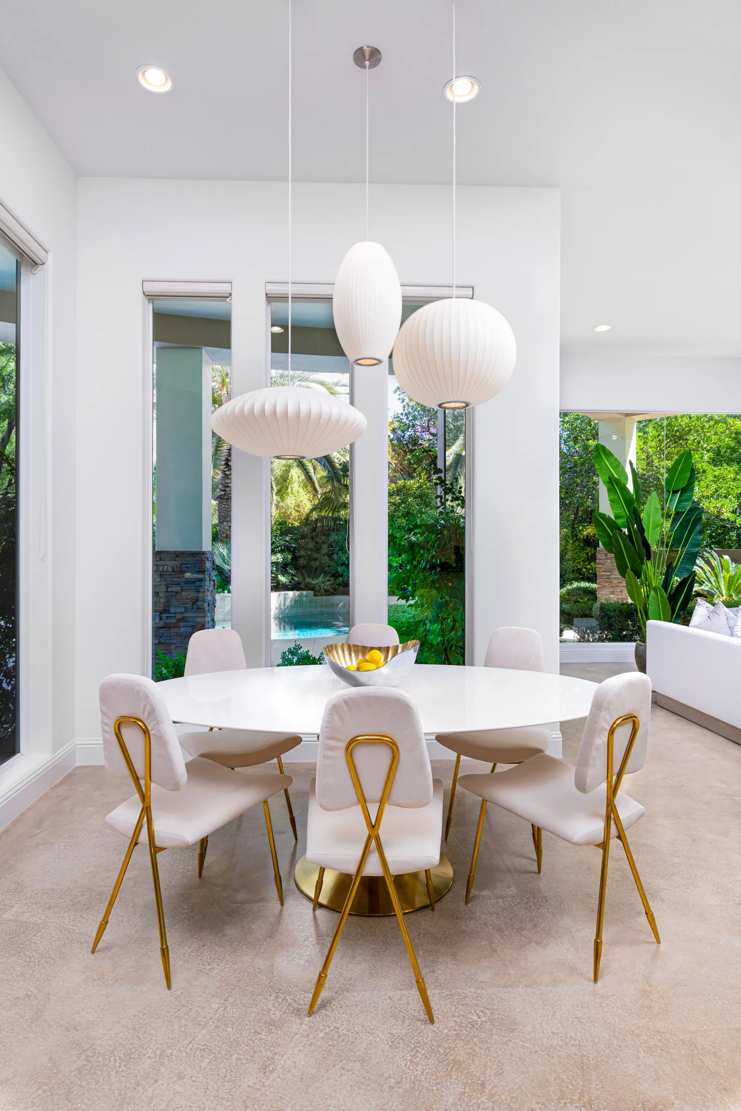 A bright dining nook with a round white table, gold-accented chairs, and large windows overlooking lush greenery and a pool.
