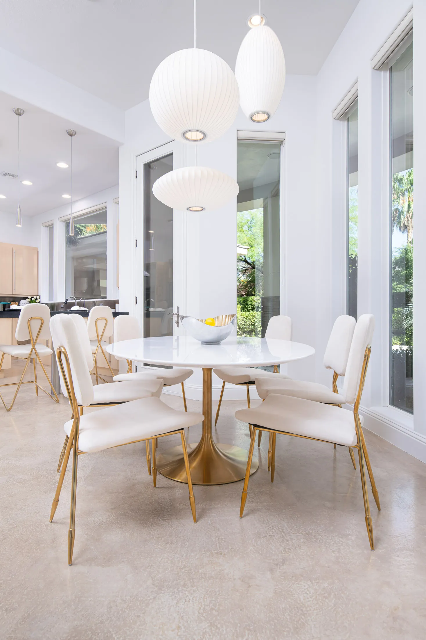 A modern dining nook featuring a white round table, gold-base chairs, and pendant lighting, with large windows bringing in natural light.