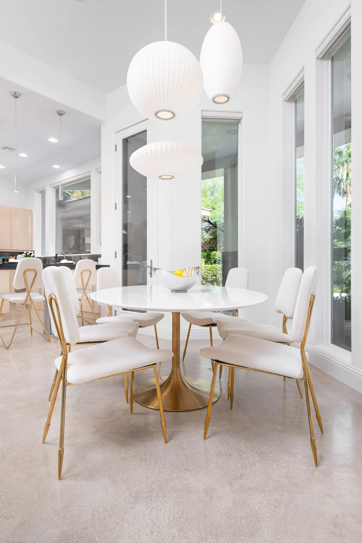 A bright dining nook with a white round table, gold-accented chairs, and large windows bringing in natural light.
