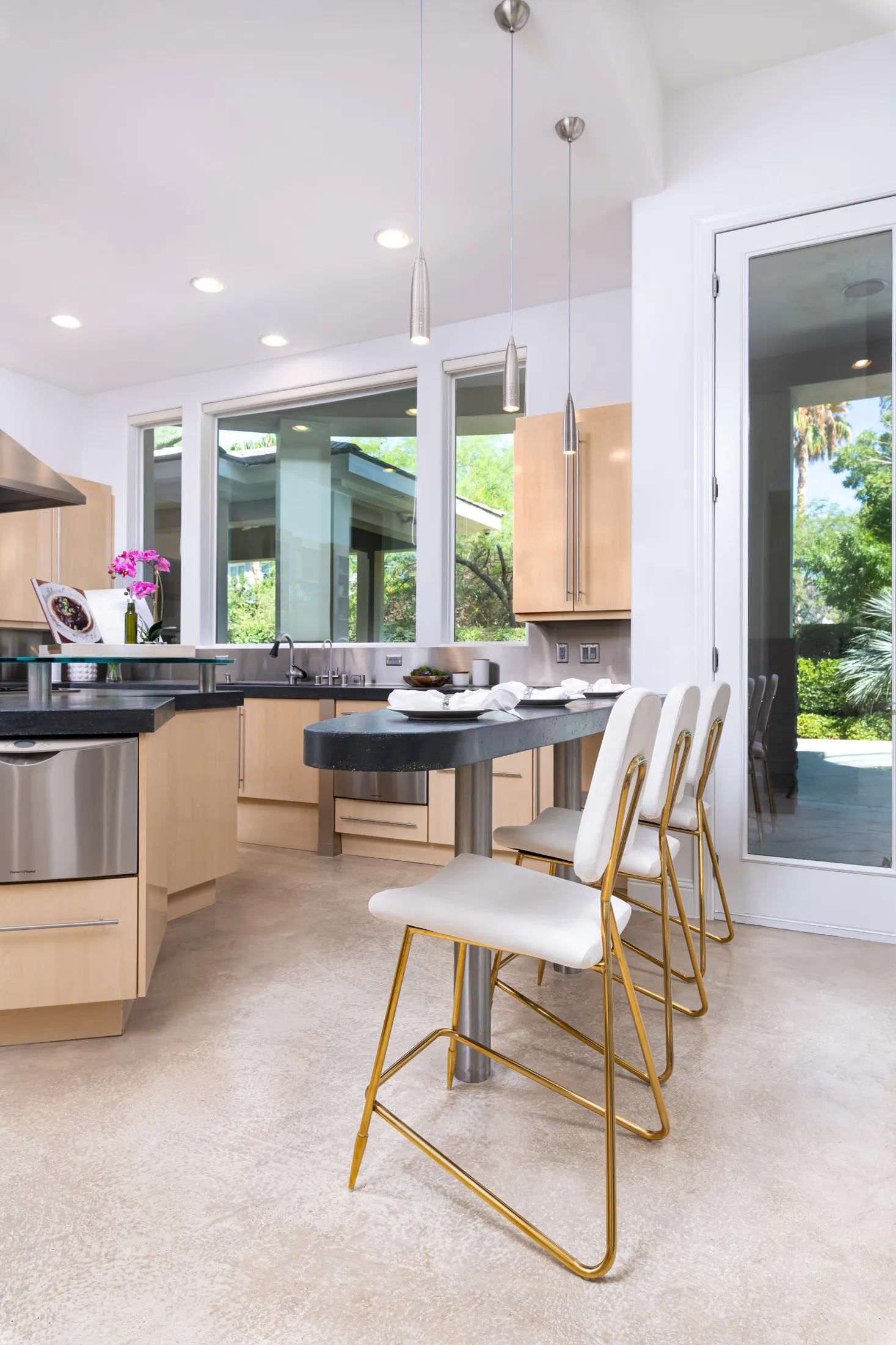 A stylish kitchen with light wood cabinetry, black countertops, white and gold barstools, and large windows overlooking lush greenery.